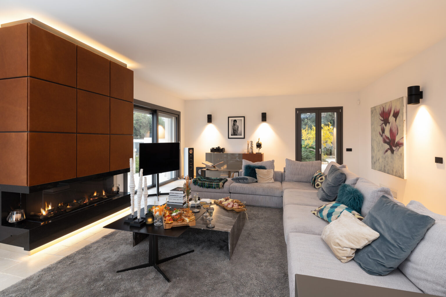 Modern living room featuring a cozy sectional sofa, a sleek coffee table with snacks, and a stylish fireplace. Natural light flows through large windows, highlighting contemporary decor and warm tones. Ideal for relaxation and social gatherings. | Only on SopranoVillas