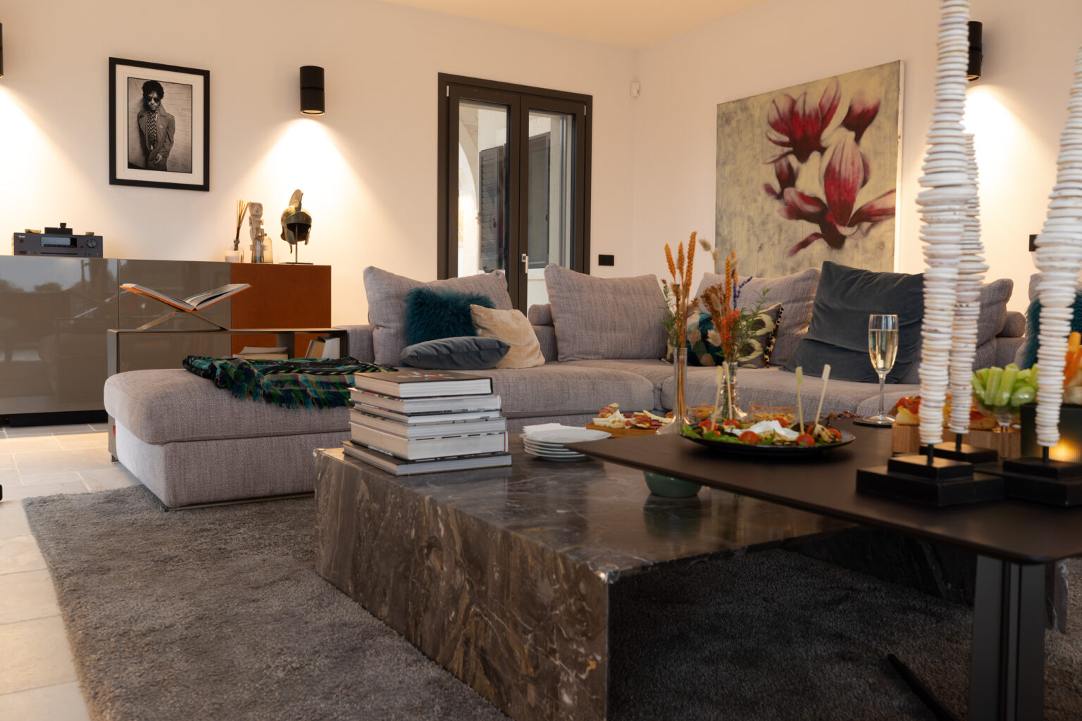 Modern living room featuring a plush gray sectional sofa with decorative pillows, a stylish marble coffee table adorned with appetizers and drinks, and artistic decor including a framed photograph and floral artwork. The space is illuminated by wall sconces, creating a cozy ambiance. | Only on SopranoVillas