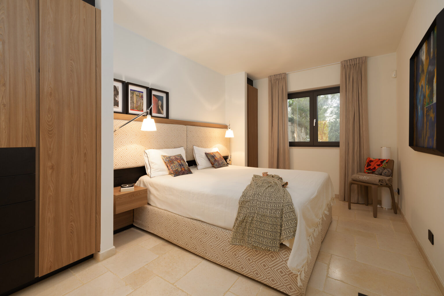 Modern bedroom interior featuring a double bed with decorative pillows, bedside lamps, and a stylish patterned bedspread. The room includes wooden cabinetry, a large window with sheer curtains, and artwork on the walls, providing a cozy and inviting atmosphere. | Only on SopranoVillas