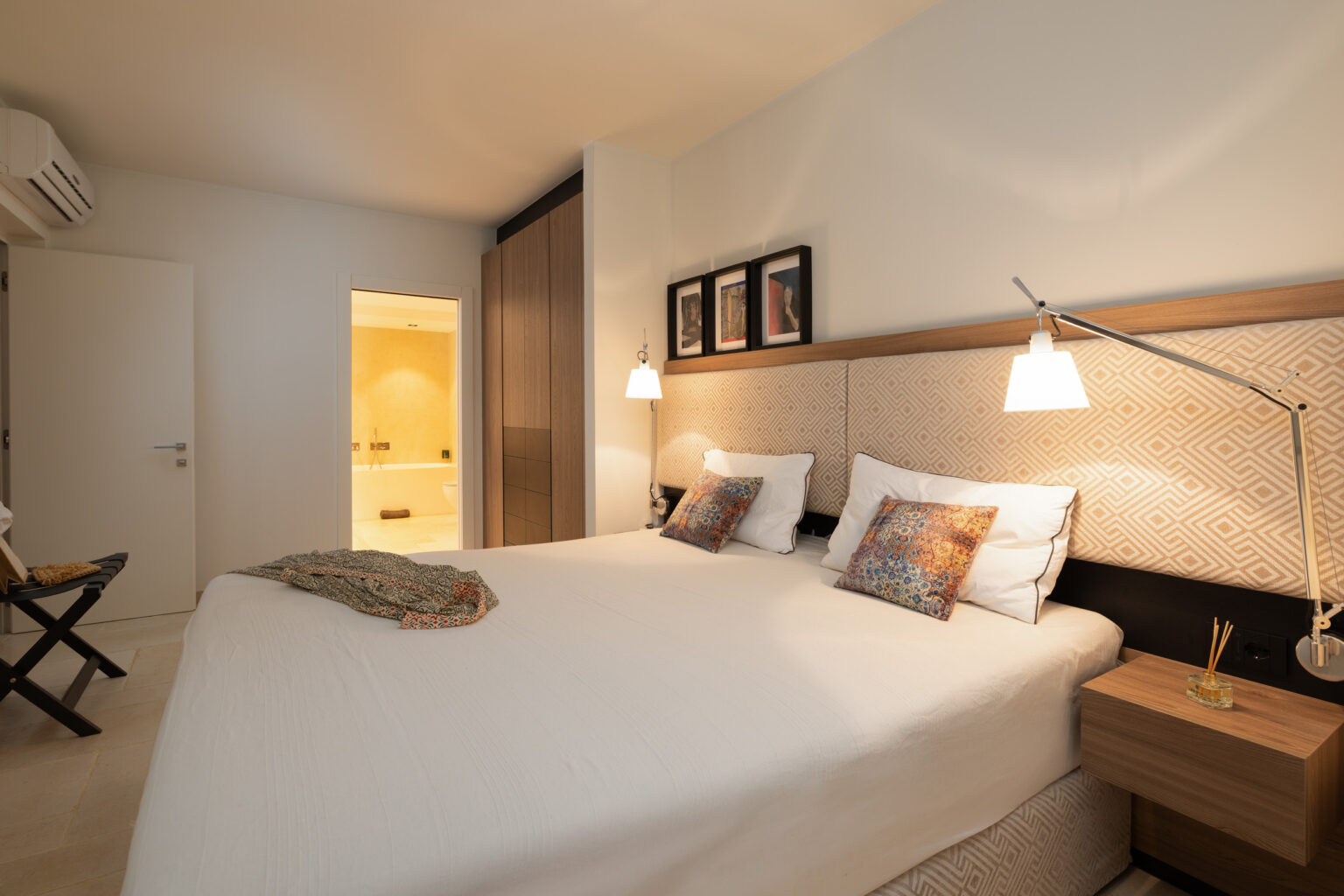 Modern hotel room featuring a double bed with decorative pillows, bedside lamps, and stylish wall art. The room includes a wooden nightstand and an open door leading to a bathroom area, creating a spacious and inviting atmosphere. | Only on SopranoVillas