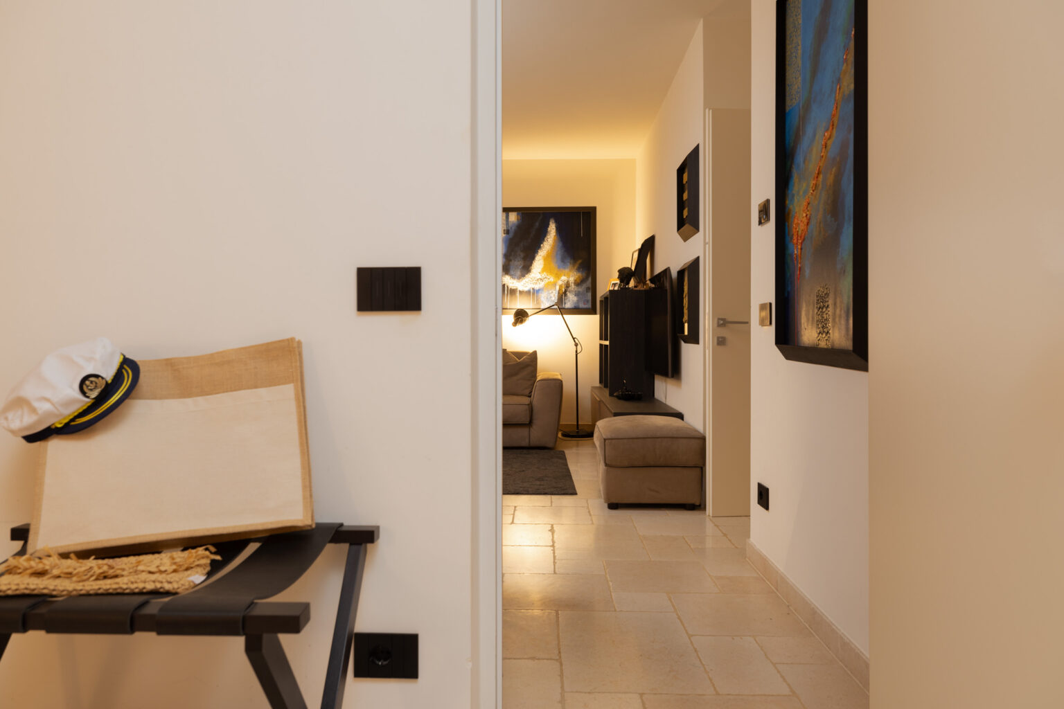 Interior view of a modern hallway leading to a cozy living room, featuring artwork on the walls, a comfortable sofa, and warm lighting. In the foreground, a folding chair and a wooden board are visible, adding to the contemporary decor. | Only on SopranoVillas