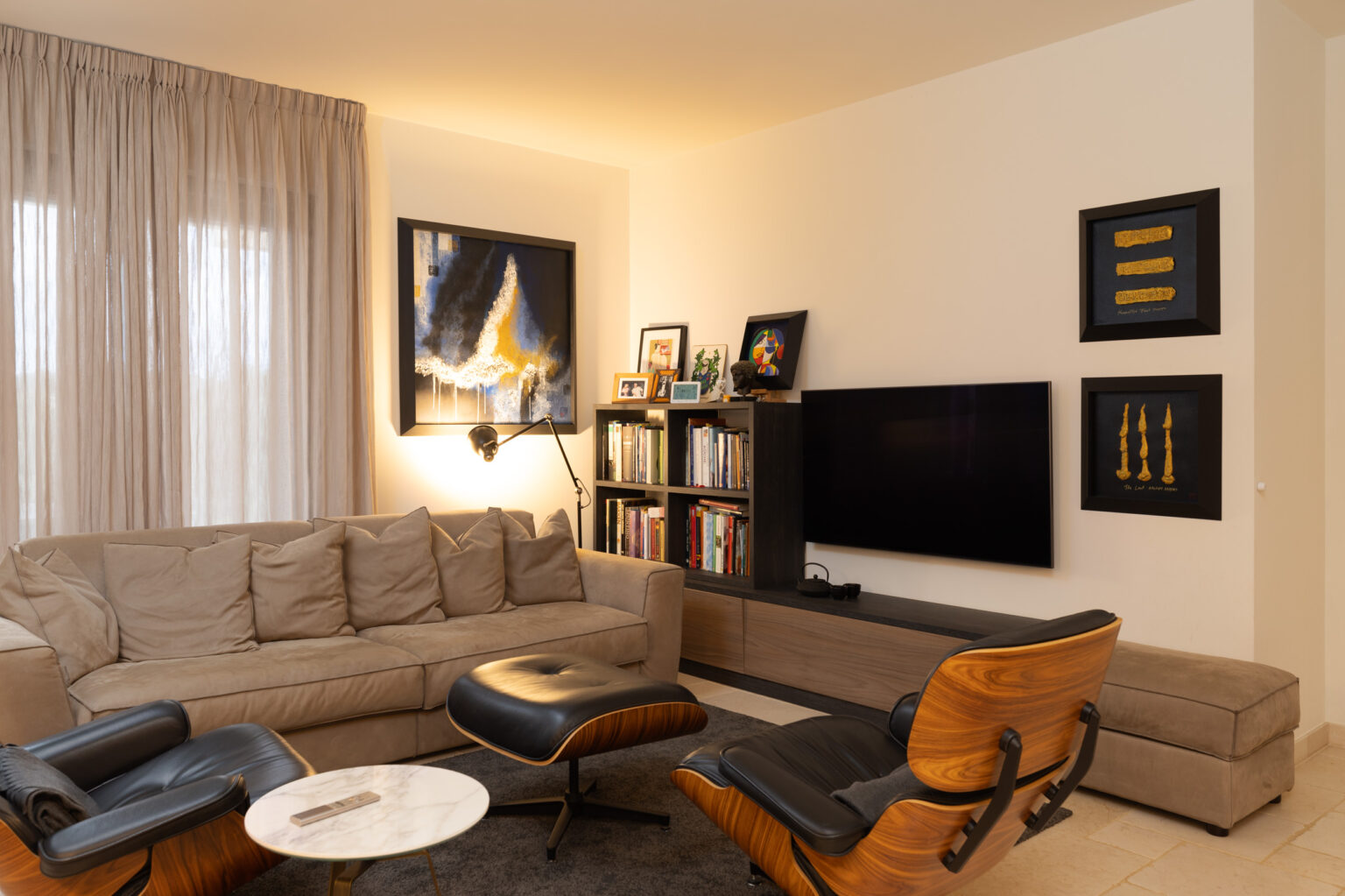 Modern living room featuring a beige sectional sofa, stylish armchairs, a flat-screen TV, and decorative artwork on the walls. The space is complemented by natural light from large windows and elegant curtains, creating a cozy and inviting atmosphere. | Only on SopranoVillas