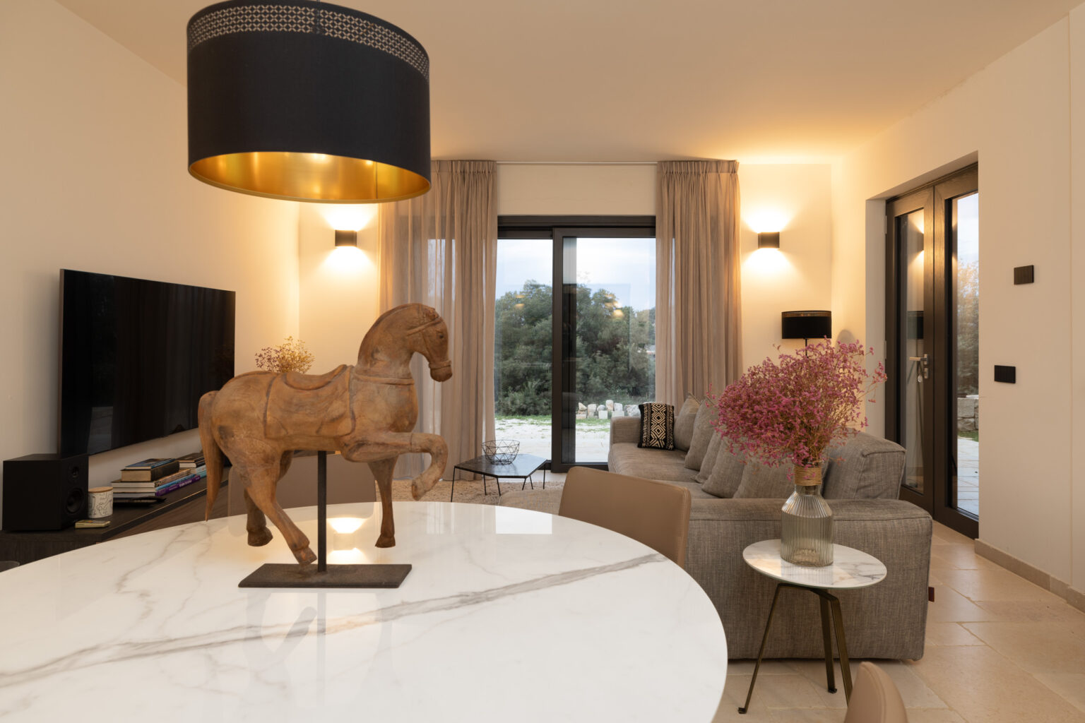 Interior view of a stylish living room featuring a decorative wooden horse statue on a marble dining table, a gray sofa with decorative cushions, and large windows with sheer curtains allowing natural light. The space includes a modern TV setup and a small side table with a vase of pink flowers, creating a warm and inviting atmosphere. | Only on SopranoVillas