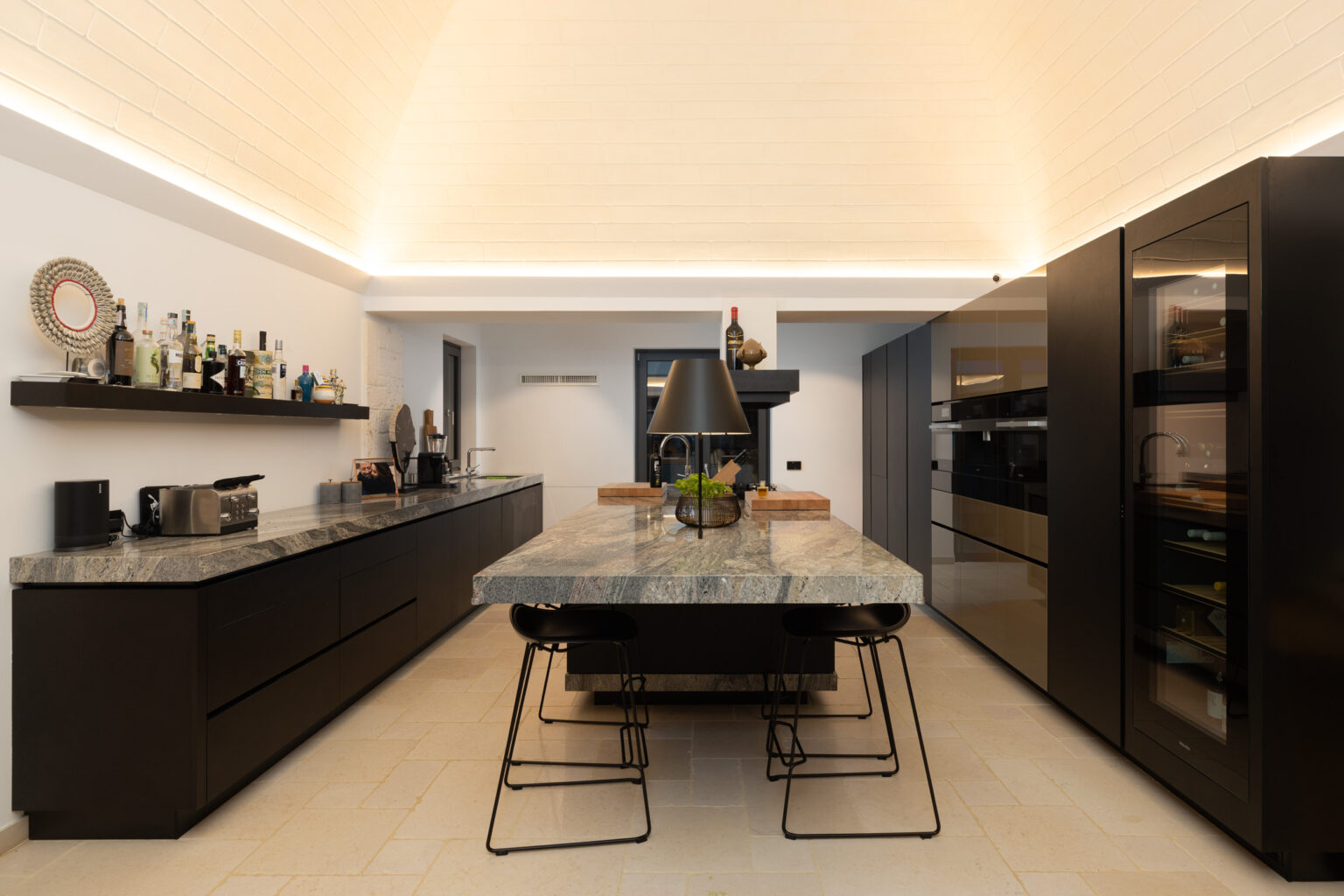 Modern kitchen interior featuring a sleek black cabinetry design, a large granite island with seating, and stylish lighting. The space is complemented by a minimalist aesthetic and organized shelves displaying various bottles and kitchen appliances. | Only on SopranoVillas