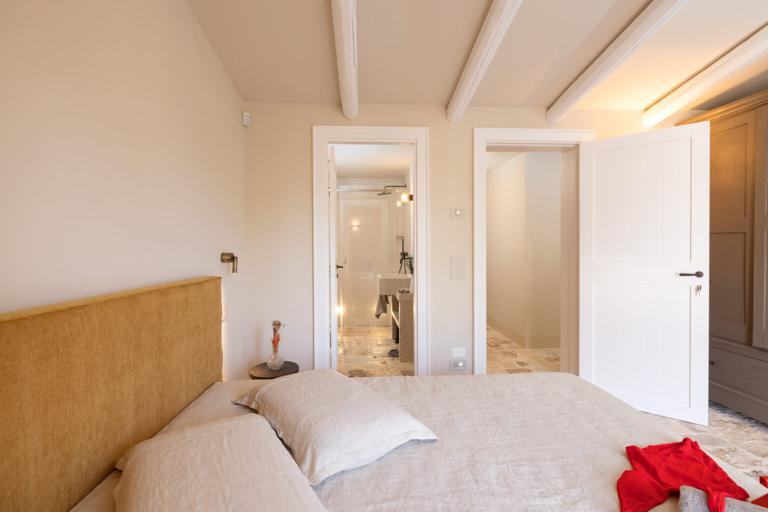 Modern bedroom interior featuring a yellow upholstered headboard, soft bedding, and a cozy atmosphere. The image reveals two doorways leading to a stylish bathroom with a glass shower and contemporary fixtures. Natural light fills the space, enhancing the warm color palette. | Only on SopranoVillas