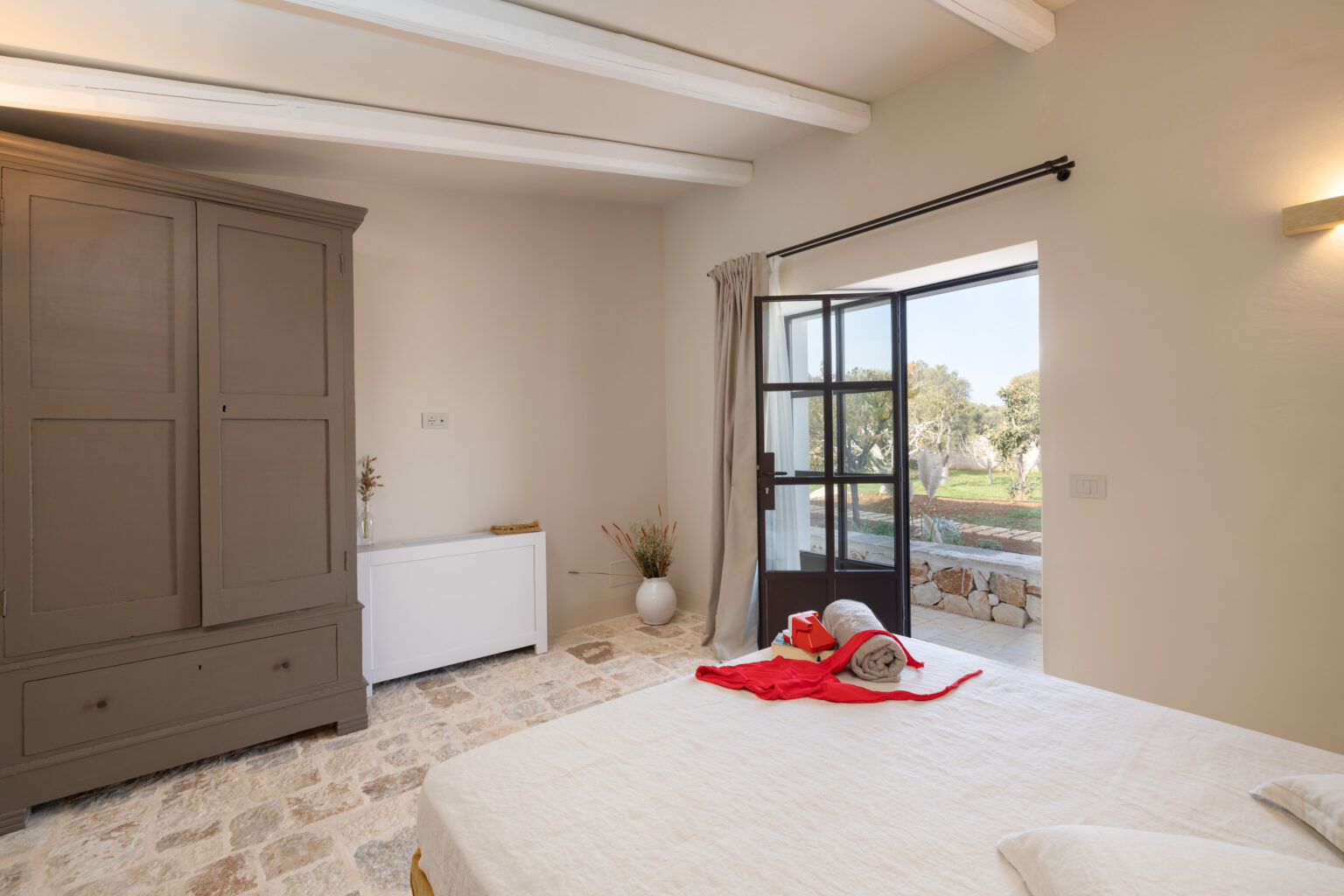 Cozy bedroom interior featuring a large wooden wardrobe, a white sideboard, and a bed with neutral bedding. Natural light floods the room through an open door leading to a garden view, complemented by decorative elements like a vase and rolled towels. | Only on SopranoVillas