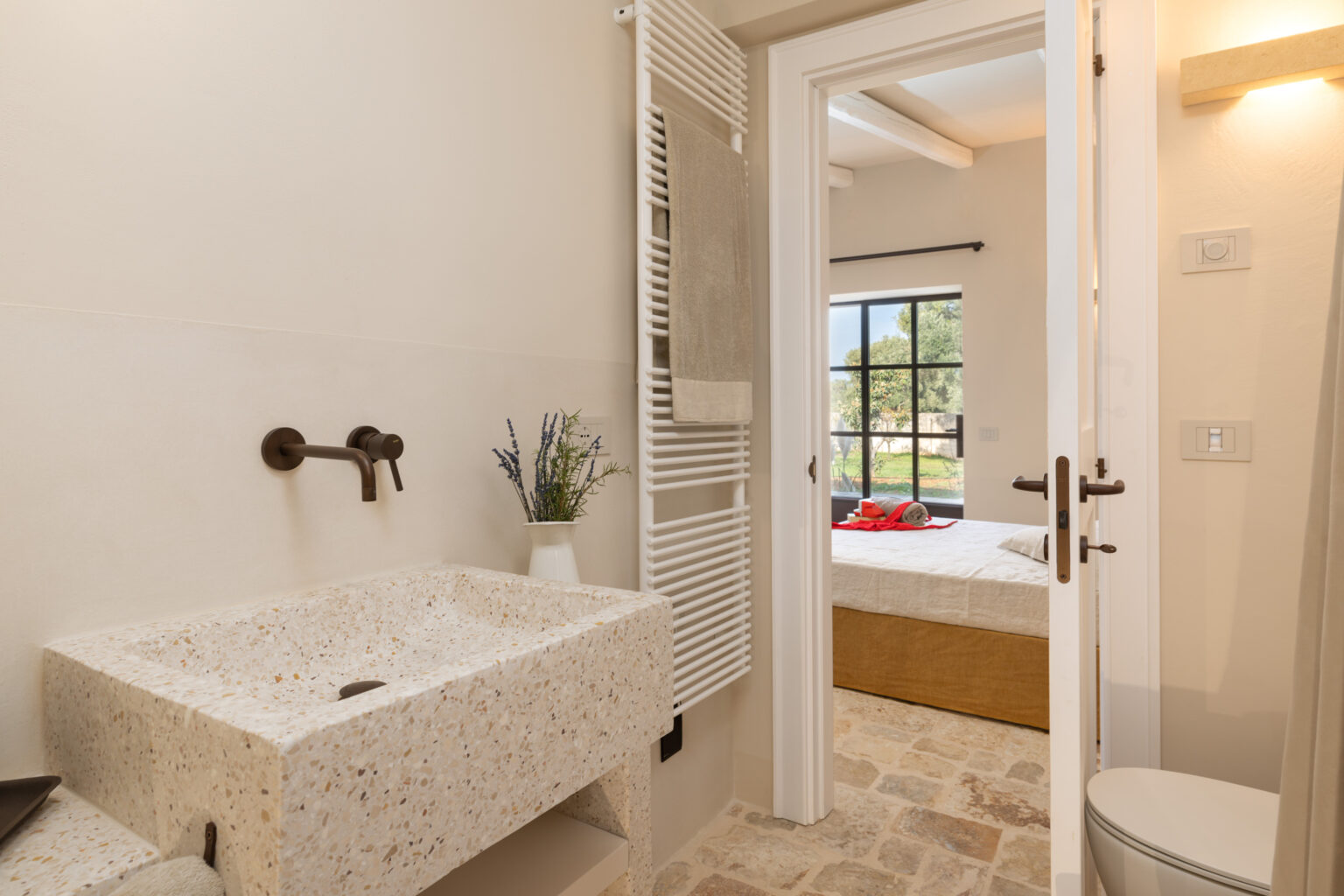 Modern bathroom featuring a stylish terrazzo sink, bronze faucets, and a towel radiator, with a view of a cozy bedroom through an open door. | Only on SopranoVillas