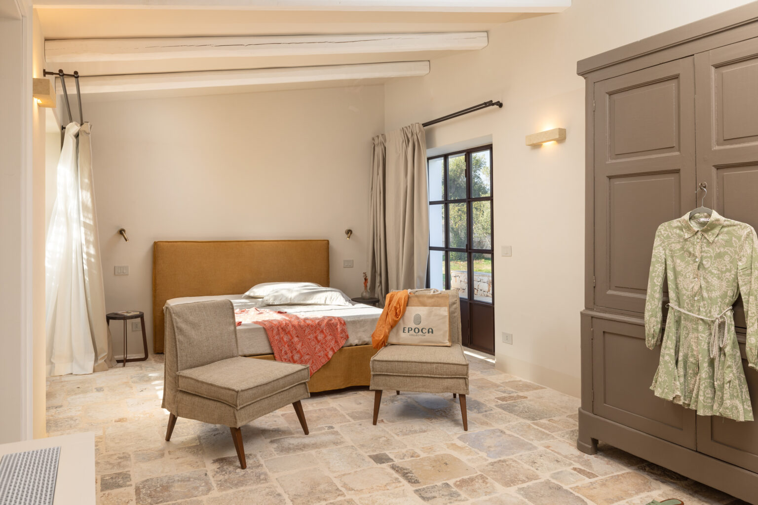 Cozy bedroom interior featuring a warm-toned bed with a decorative throw, two stylish chairs, and a vintage wardrobe. Natural light streams through a large window, highlighting the stone flooring and soft color palette, creating a serene and inviting atmosphere. | Only on SopranoVillas