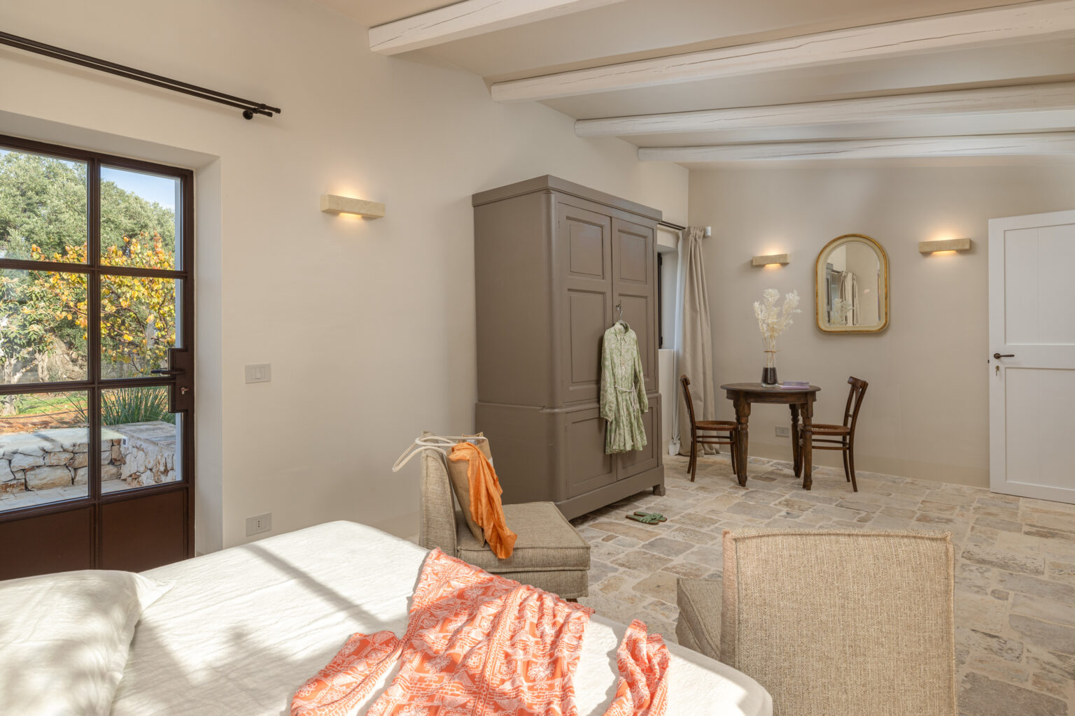 Cozy and stylish interior of a bedroom featuring a bed with white linens, a beige armchair, a wooden wardrobe, and a small dining table with chairs. Natural light floods the room through a large window, highlighting the stone flooring and decor elements like a mirror and floral arrangement. | Only on SopranoVillas