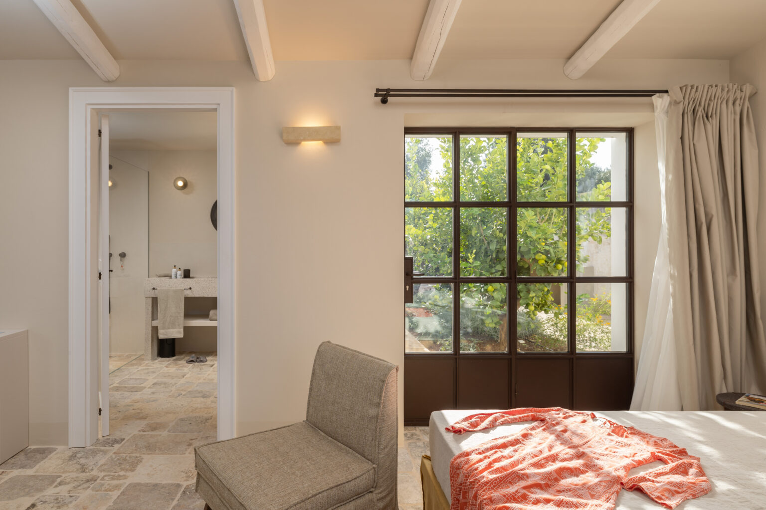 Bright and airy bedroom featuring a cozy chair, a bed with a patterned throw, and a view of lush greenery through large windows. The room includes a doorway leading to a modern bathroom with stone accents and natural light. Ideal for showcasing contemporary interior design and tranquil living spaces. | Only on SopranoVillas