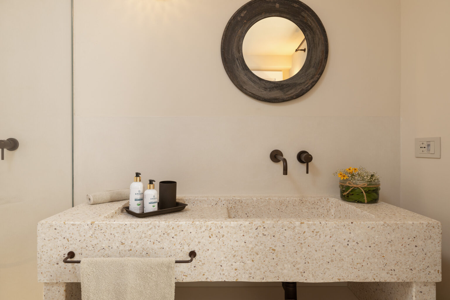 Modern bathroom featuring a minimalist stone sink with sleek black faucets, a round mirror, and a decorative tray holding shampoo and conditioner bottles, complemented by a small plant arrangement. | Only on SopranoVillas