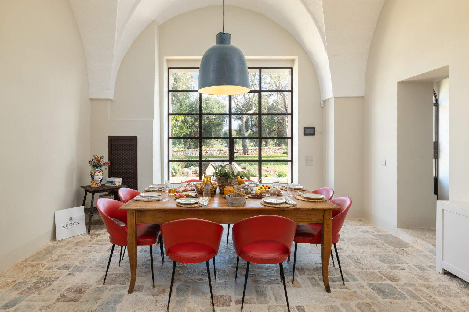 Bright and spacious dining room featuring a wooden table set for a meal, surrounded by red chairs. Large windows provide natural light and a view of the garden, while a modern pendant light hangs overhead. Decorative elements include a colorful vase and a stylish sign on the floor. | Only on SopranoVillas