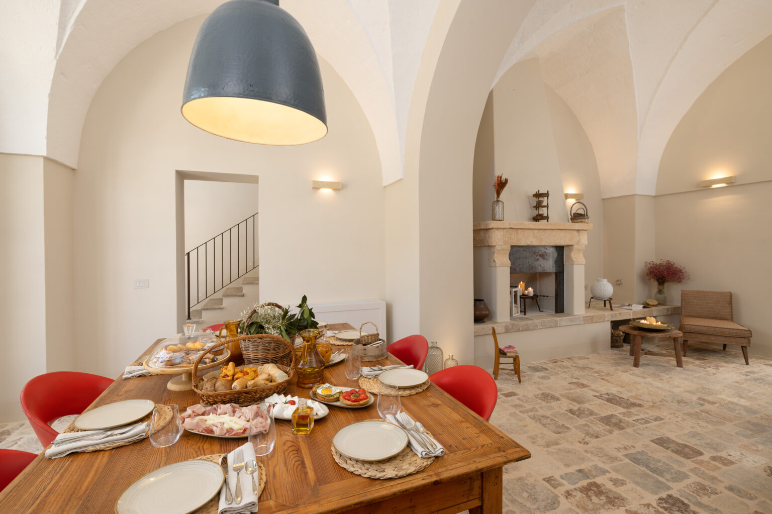 Elegant dining area featuring a rustic wooden table set with plates and glassware, surrounded by red chairs. A variety of gourmet foods, including meats and pastries, are displayed on the table. The space is illuminated by a large pendant light, with an arched ceiling and a cozy fireplace in the background, creating a warm and inviting atmosphere. | Only on SopranoVillas
