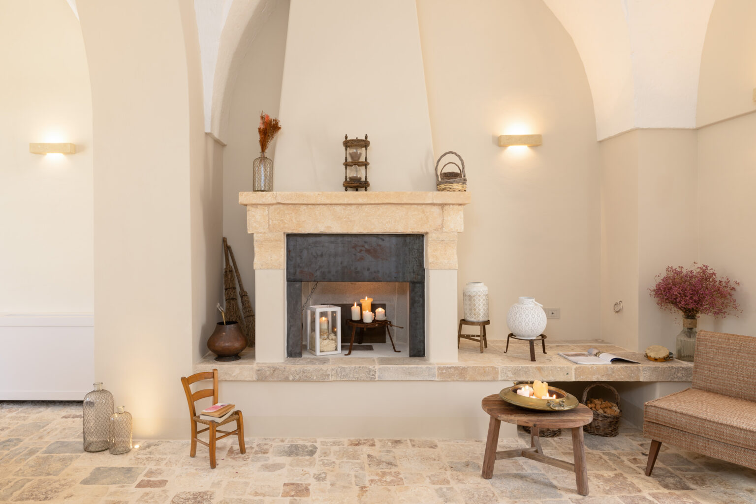 Cozy interior featuring a stone fireplace adorned with candles, decorative vases, and a small wooden chair. The space includes a rustic table with a bowl of snacks, a comfortable seating area, and warm lighting, creating an inviting atmosphere. Ideal for home decor inspiration. | Only on SopranoVillas