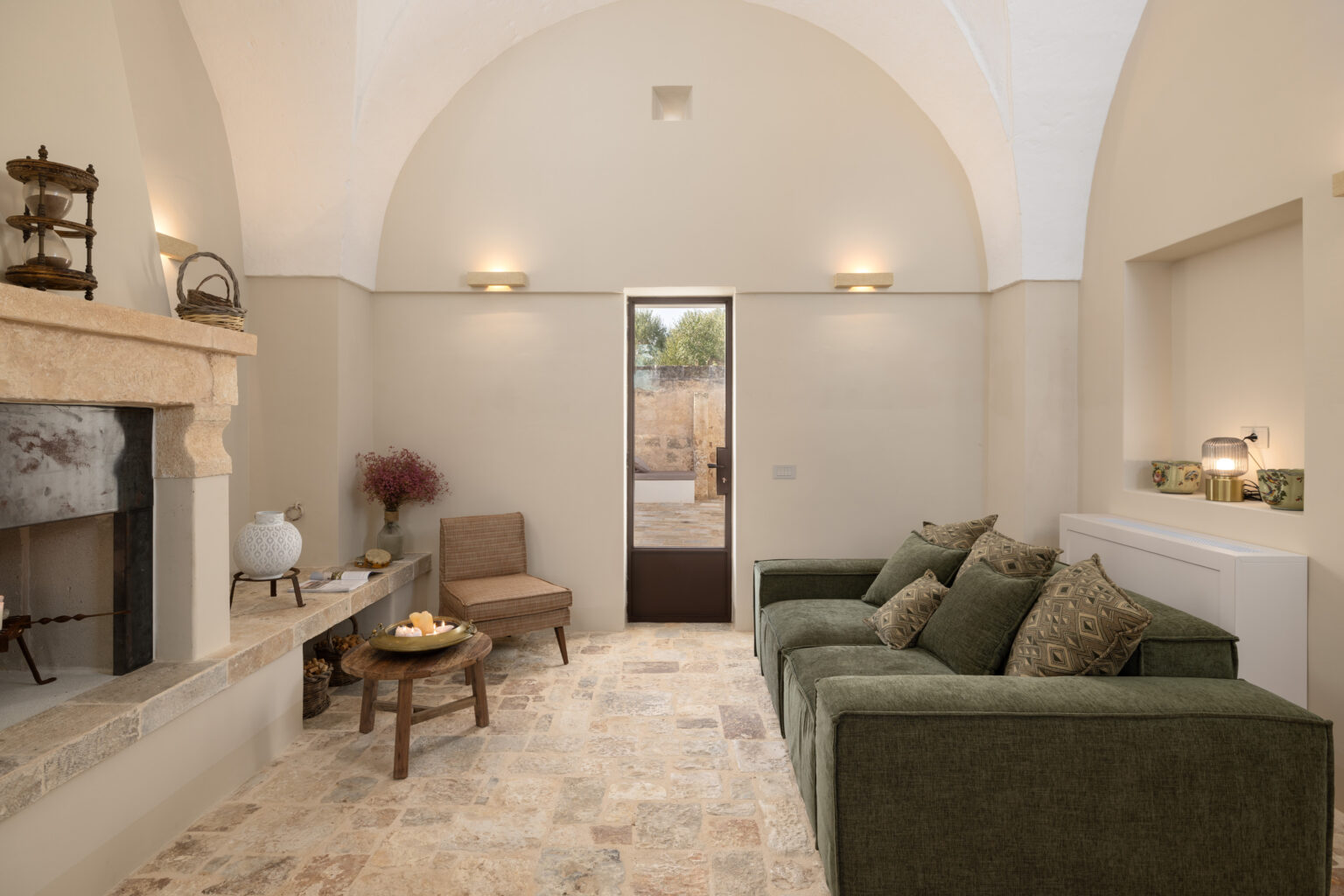 Cozy living room featuring a green sofa with decorative pillows, a stone fireplace, and a wooden coffee table. The room has high arched ceilings, warm lighting, and a door leading to an outdoor space with natural views. | Only on SopranoVillas