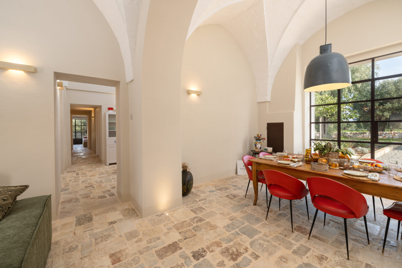 Modern dining area featuring a wooden table surrounded by red chairs, stone flooring, and large windows with a view of greenery. The space includes an arched ceiling, decorative elements, and a warm, inviting atmosphere. Ideal for family gatherings or entertaining guests. | Only on SopranoVillas