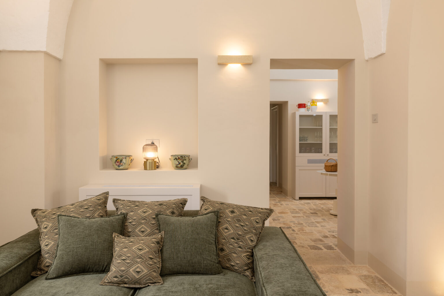 Cozy living room featuring a green sofa adorned with patterned cushions, a decorative lamp on a shelf, and glimpses of a well-lit kitchen area in the background with a modern design. The warm, neutral color palette and stone flooring create an inviting atmosphere. | Only on SopranoVillas