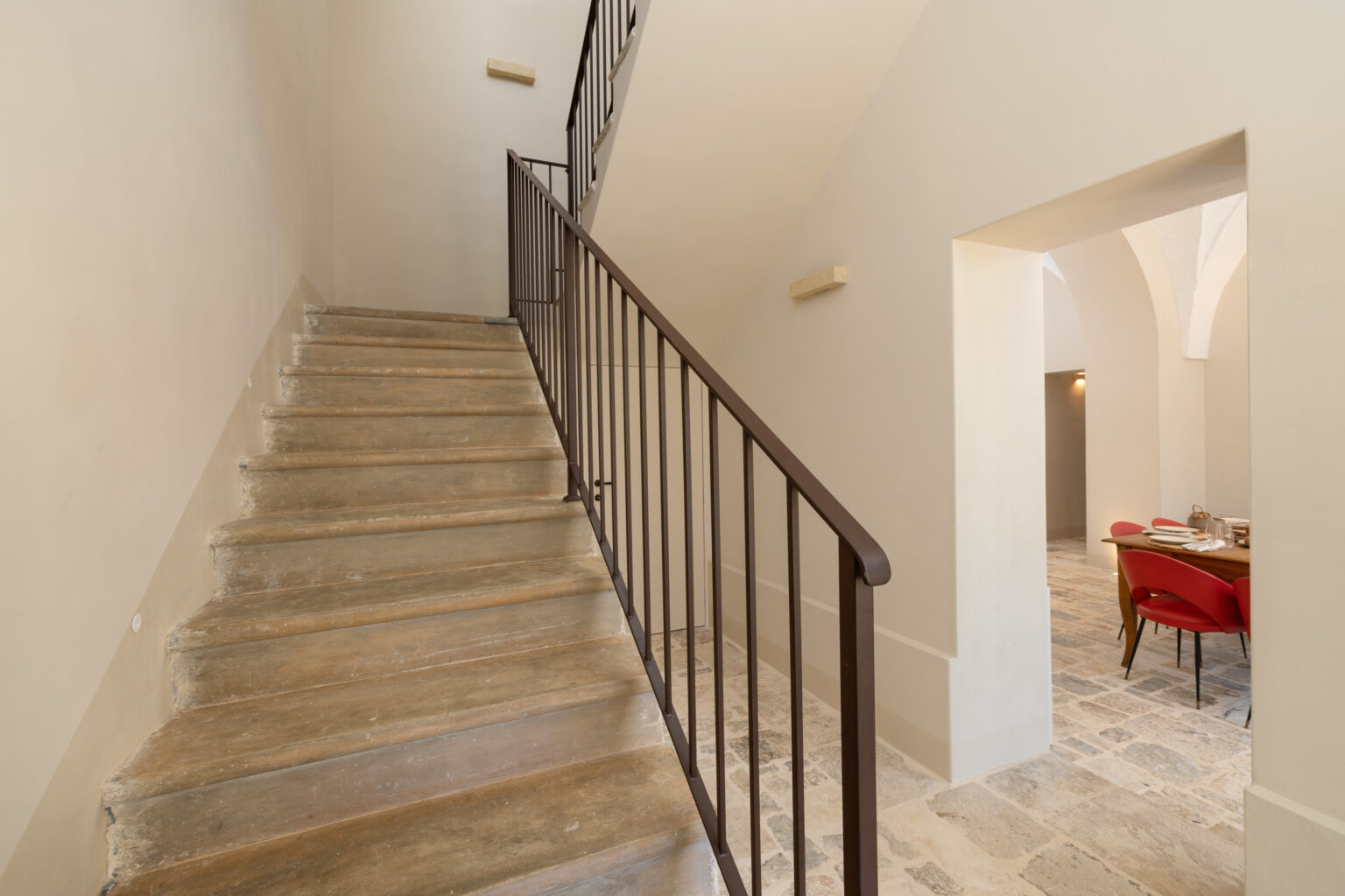 Interior view of a staircase leading to an upper level, featuring wooden steps and a black metal railing, with a glimpse of a dining area in the background. The walls are painted in a soft neutral color, enhancing the spacious and modern design. | Only on SopranoVillas