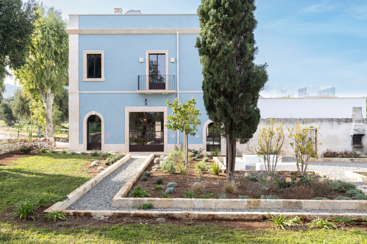 Charming blue house with stone accents surrounded by a landscaped garden featuring various plants and gravel pathways, set against a clear sky. | Only on SopranoVillas