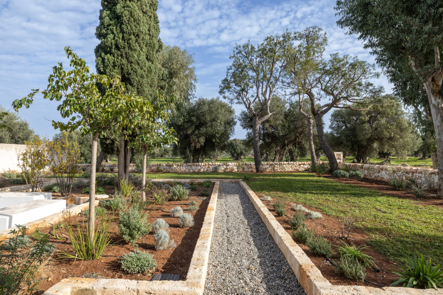 Lush garden pathway lined with greenery, featuring a mix of trees and shrubs, with a stone wall in the background under a partly cloudy sky. | Only on SopranoVillas