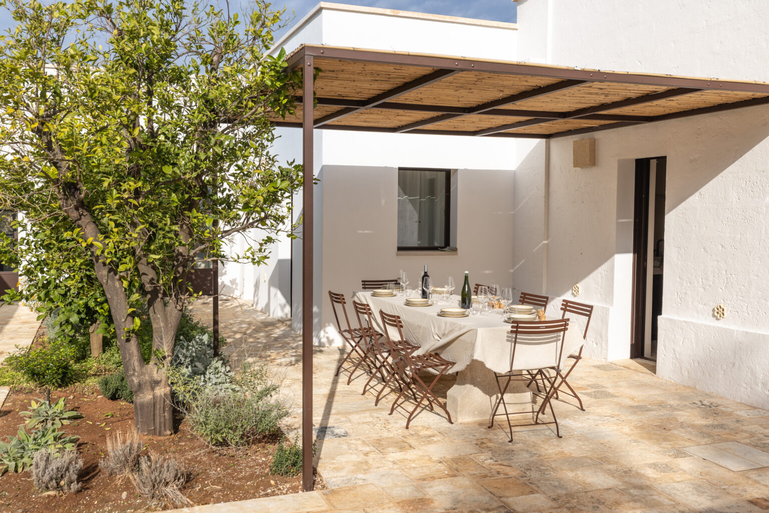 Outdoor dining area featuring a beautifully set table with plates and glasses, surrounded by greenery and a lemon tree, under a bamboo-covered pergola, ideal for al fresco meals in a sunny setting. | Only on SopranoVillas