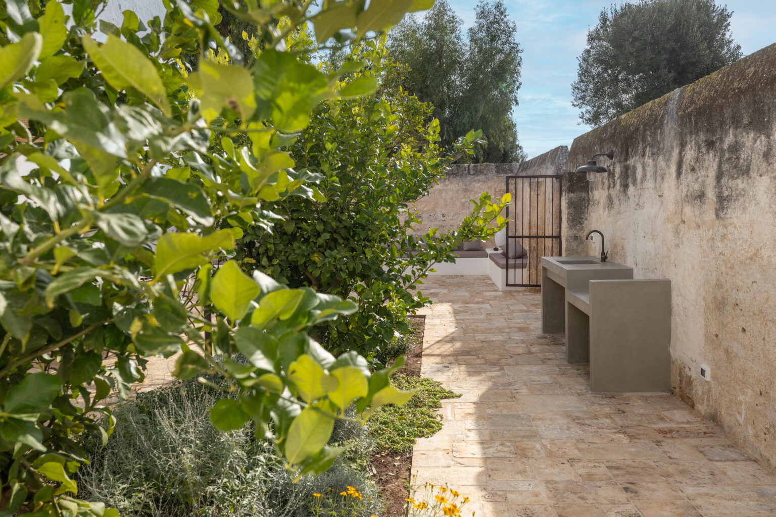Lush garden pathway featuring greenery, stone flooring, and outdoor sinks against a rustic wall. Ideal for outdoor living and gardening. | Only on SopranoVillas