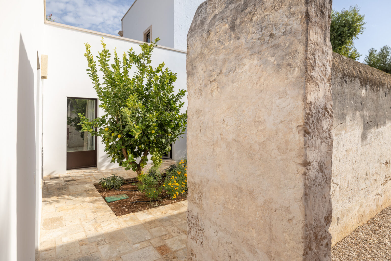 Modern courtyard featuring a vibrant lemon tree and neatly arranged flower beds, surrounded by a rustic stone wall and sleek white architecture, creating a serene outdoor space. | Only on SopranoVillas