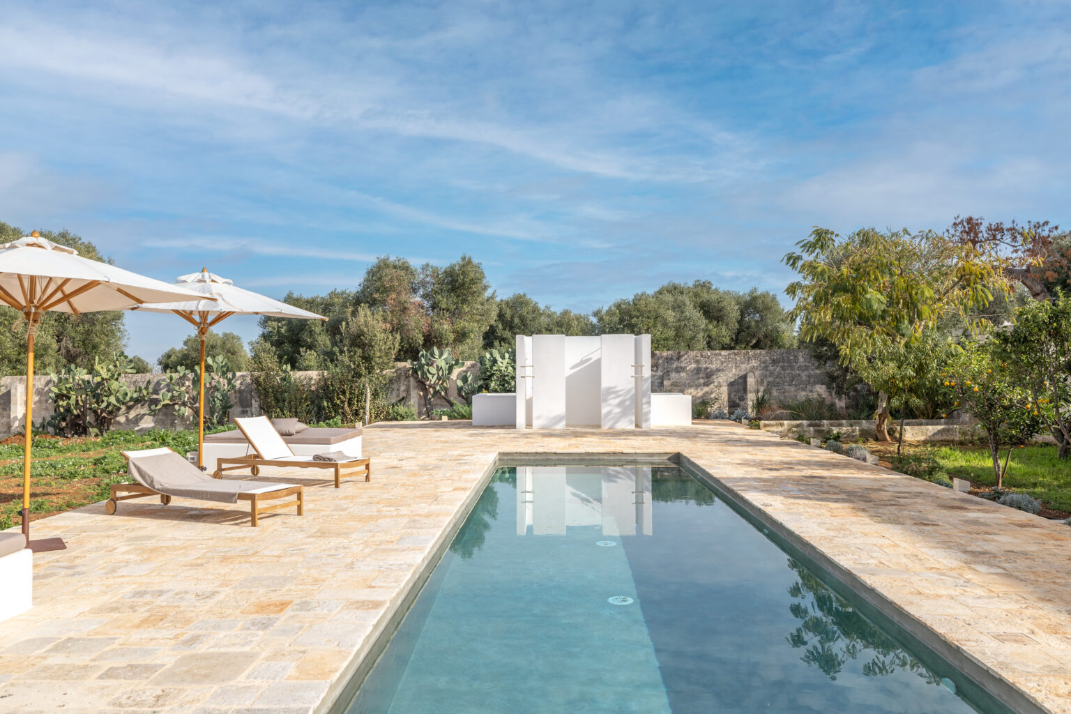 Modern outdoor pool area featuring comfortable lounge chairs and umbrellas, surrounded by lush greenery and stone walls, under a clear blue sky. Ideal for relaxation and leisure in a serene setting. | Only on SopranoVillas