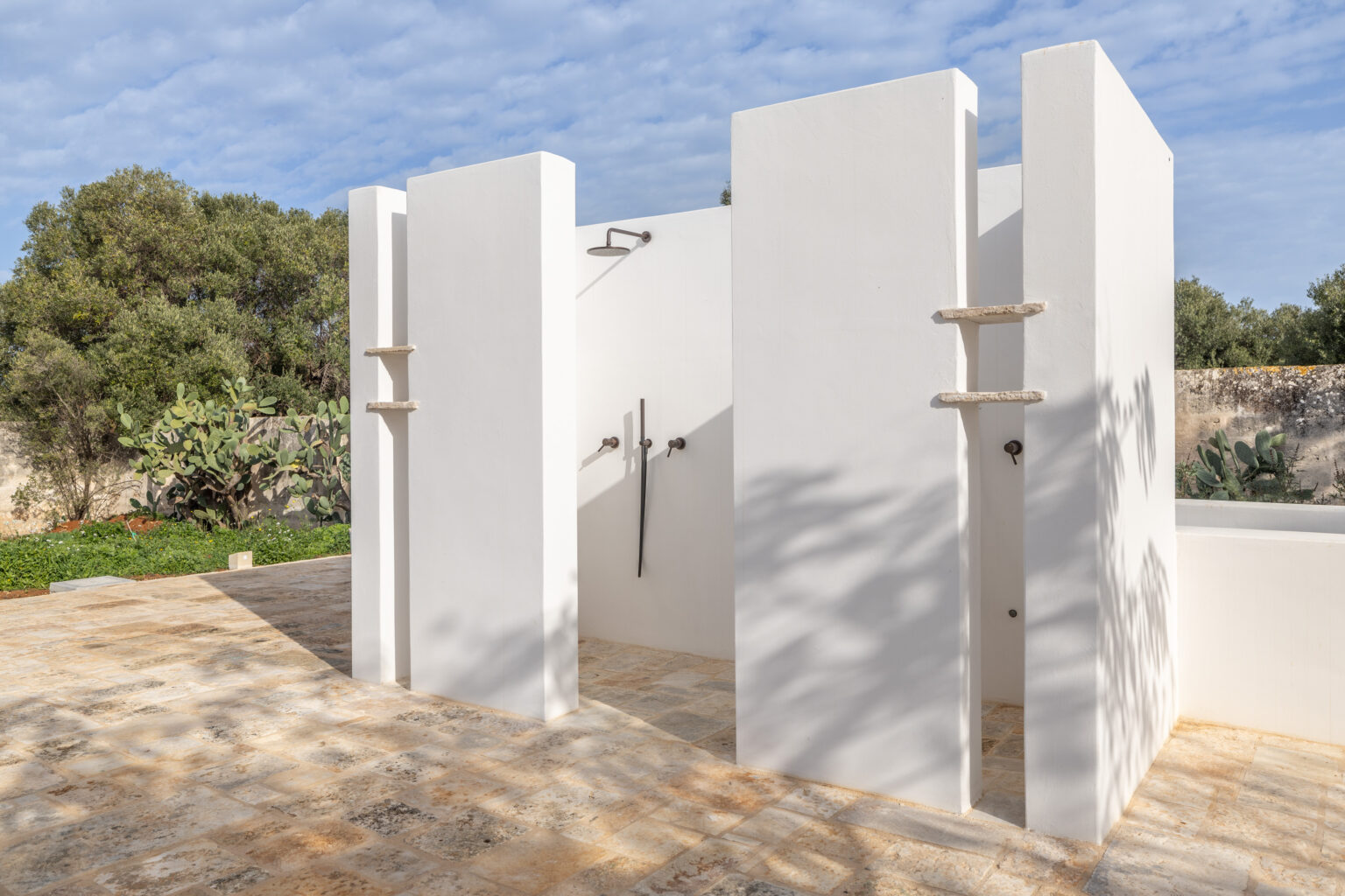 Outdoor shower area featuring minimalist white walls and modern fixtures, surrounded by lush greenery and natural stone flooring, ideal for luxury home designs and outdoor living spaces. | Only on SopranoVillas