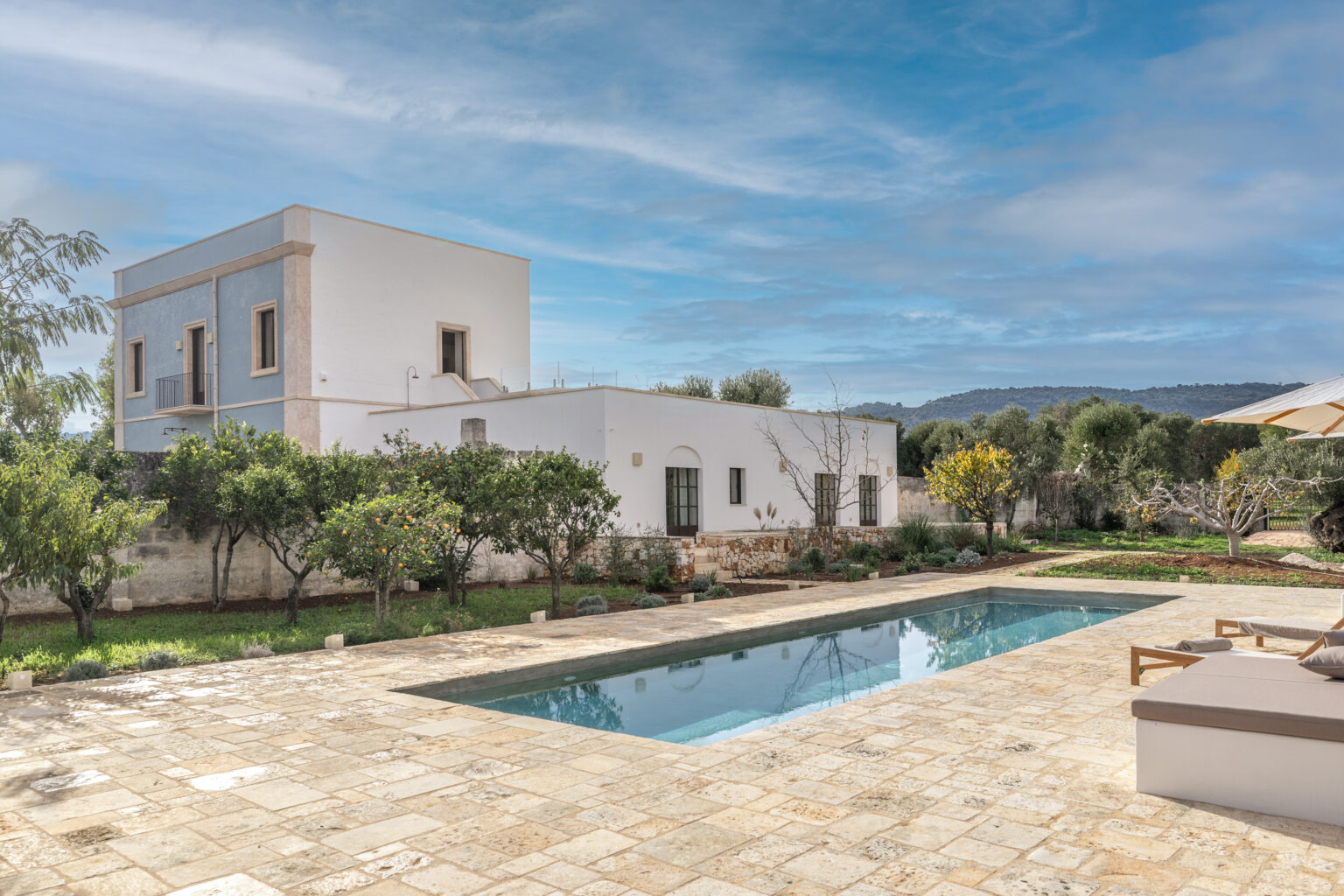 Modern villa with a swimming pool surrounded by lush greenery and olive trees, featuring a blend of traditional and contemporary architecture against a clear blue sky. Ideal for luxury retreats and outdoor relaxation. | Only on SopranoVillas