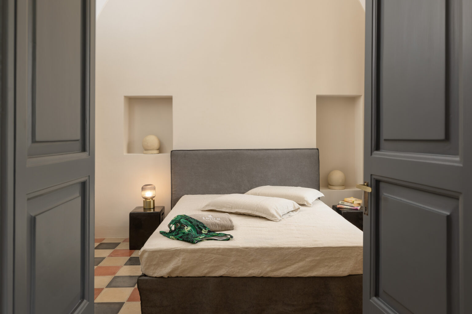 Cozy bedroom interior featuring a gray upholstered bed with white linens, decorative pillows, and a green scarf. The room has a minimalist design with soft lighting from a bedside lamp, showcasing a warm color palette of tiled flooring and neutral walls. Ideal for relaxation and comfort. | Only on SopranoVillas
