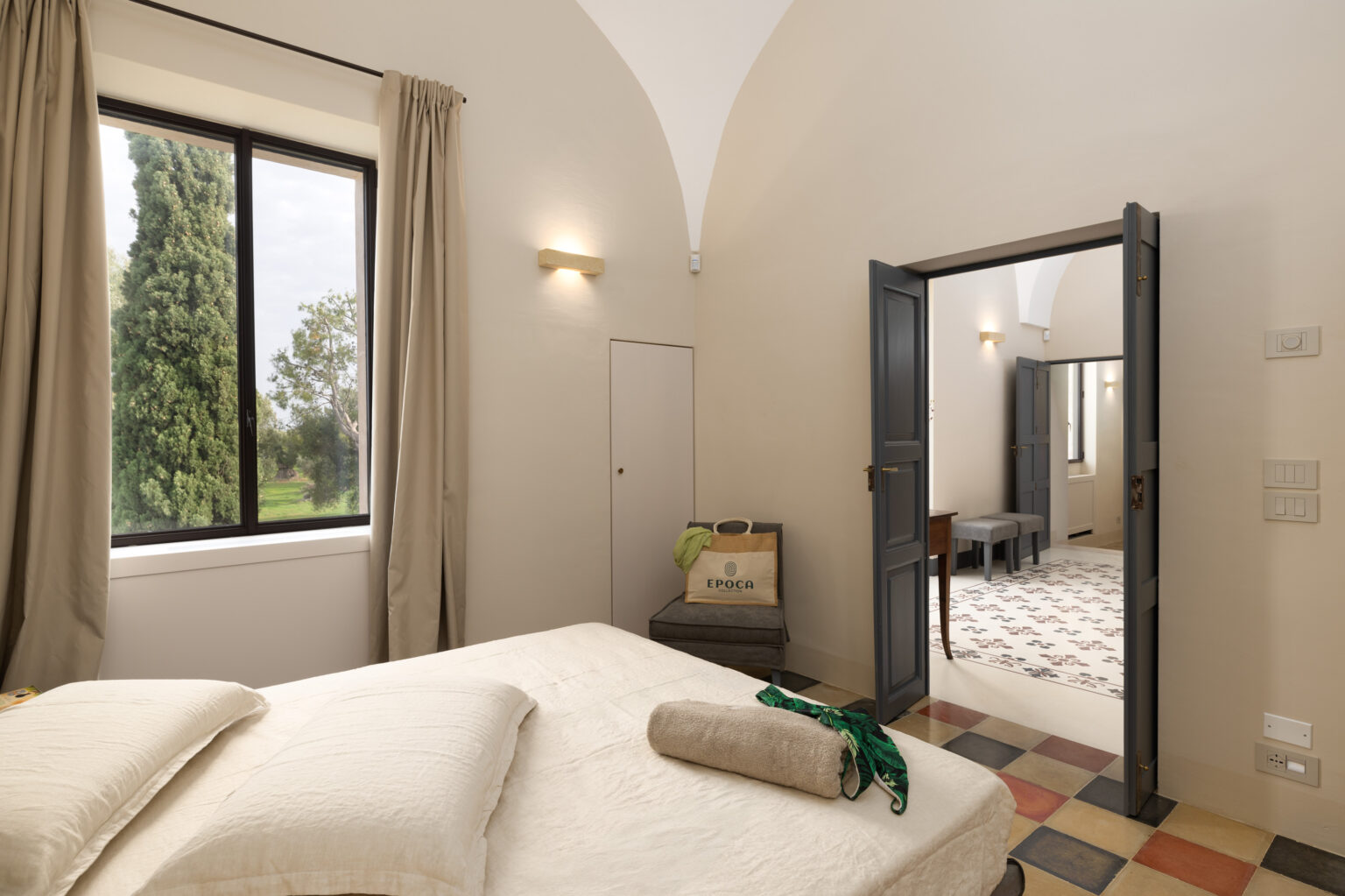 Cozy bedroom with a neutral color palette, featuring a comfortable bed, large window with natural light, and views of greenery. An open doorway leads to a stylish adjoining room with patterned flooring. | Only on SopranoVillas