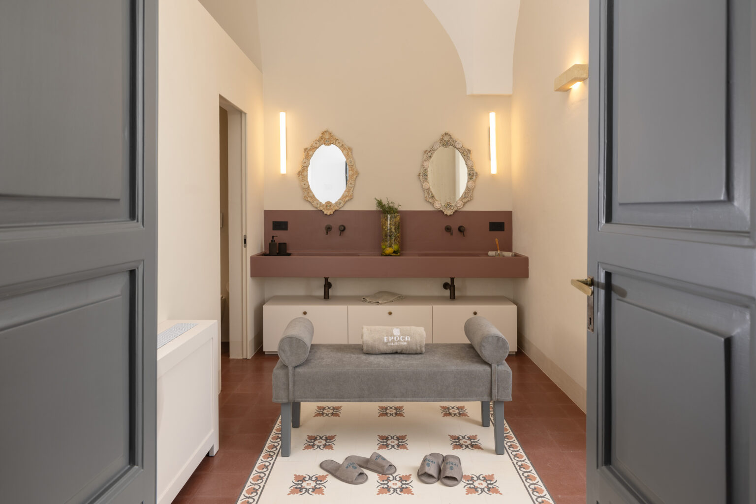 Modern bathroom interior featuring a sleek design with dual decorative mirrors, a stylish gray bench, and a patterned floor. The space includes a minimalist sink area with chic fixtures and ambient lighting, creating a serene atmosphere. Soft slippers and a towel add a touch of comfort, enhancing the elegant decor. | Only on SopranoVillas