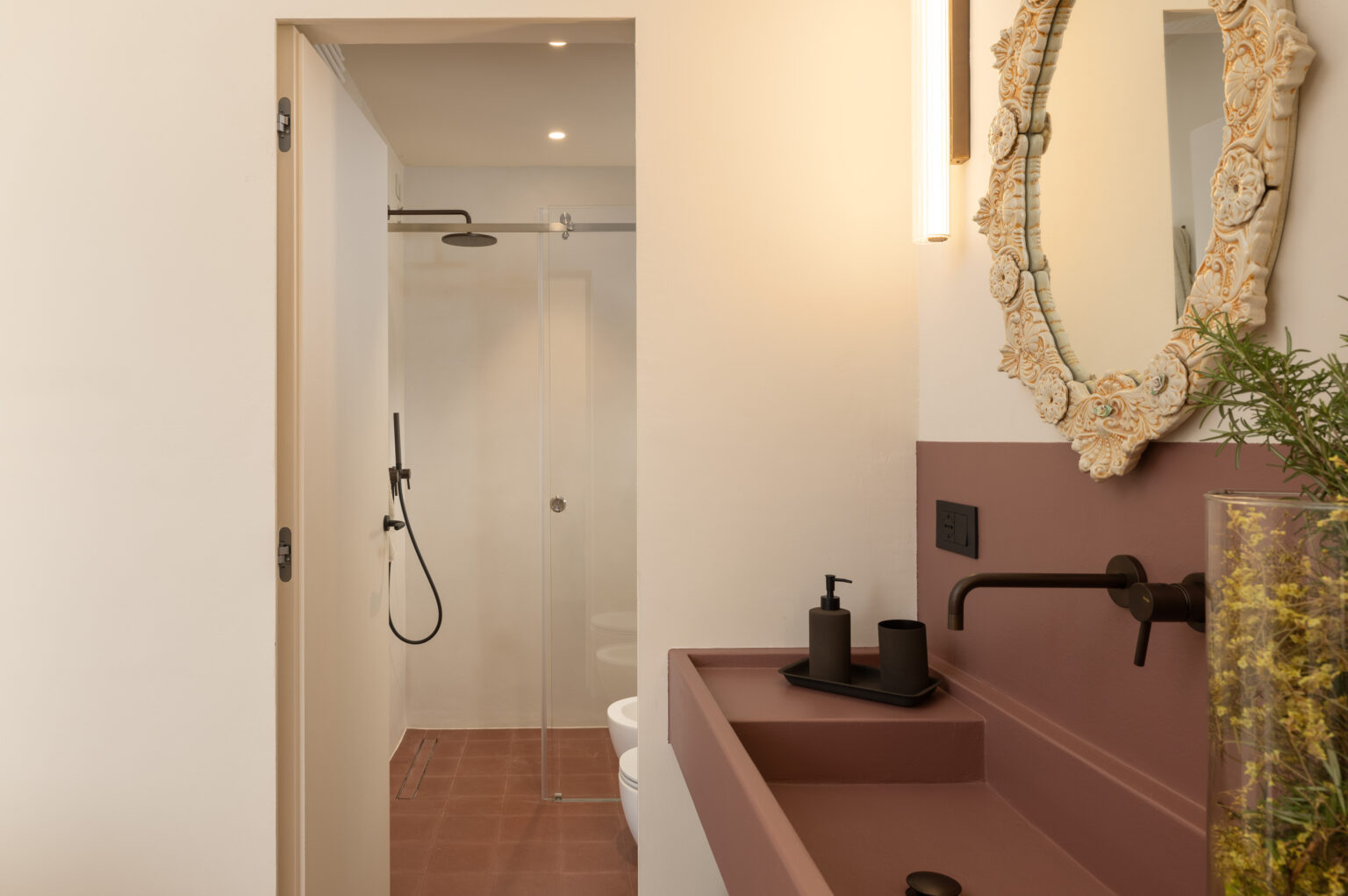 Modern bathroom interior featuring a stylish sink with black fixtures, a decorative mirror, and a glass shower enclosure. The walls are painted in soft tones, complemented by earthy terracotta flooring, creating an inviting and elegant atmosphere. | Only on SopranoVillas