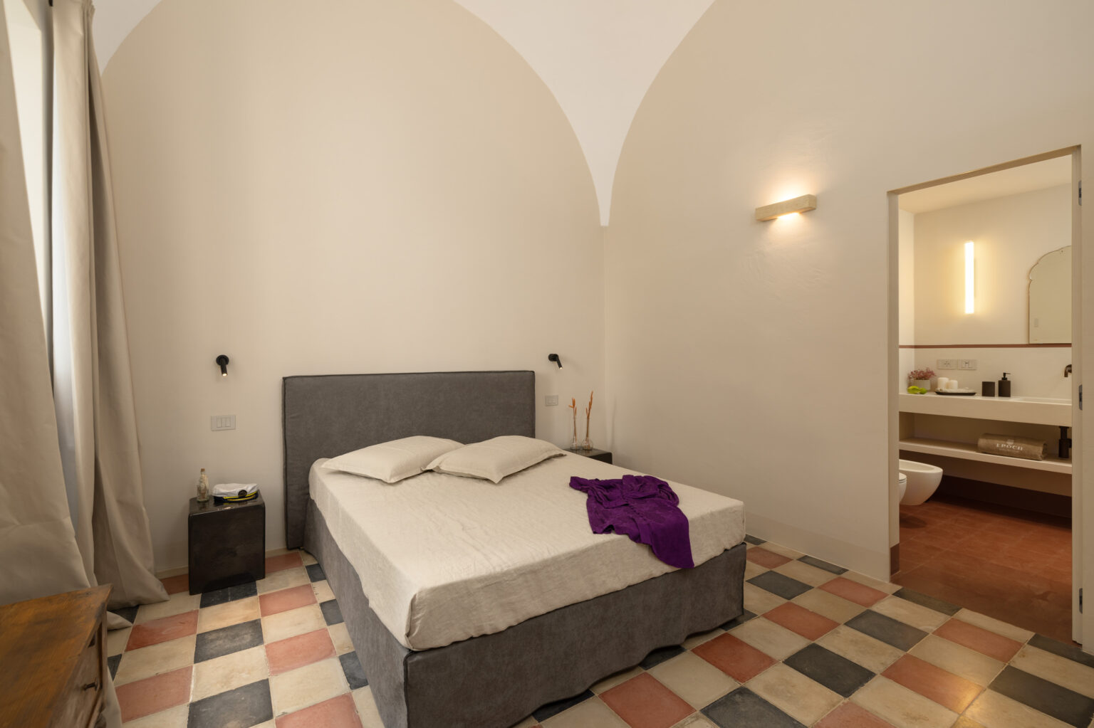 Modern bedroom featuring a gray upholstered bed with two pillows, a purple throw, and a colorful patterned tiled floor. The room has an arched ceiling, natural light from a window, and leads to a minimalist bathroom with a sink and toilet. | Only on SopranoVillas