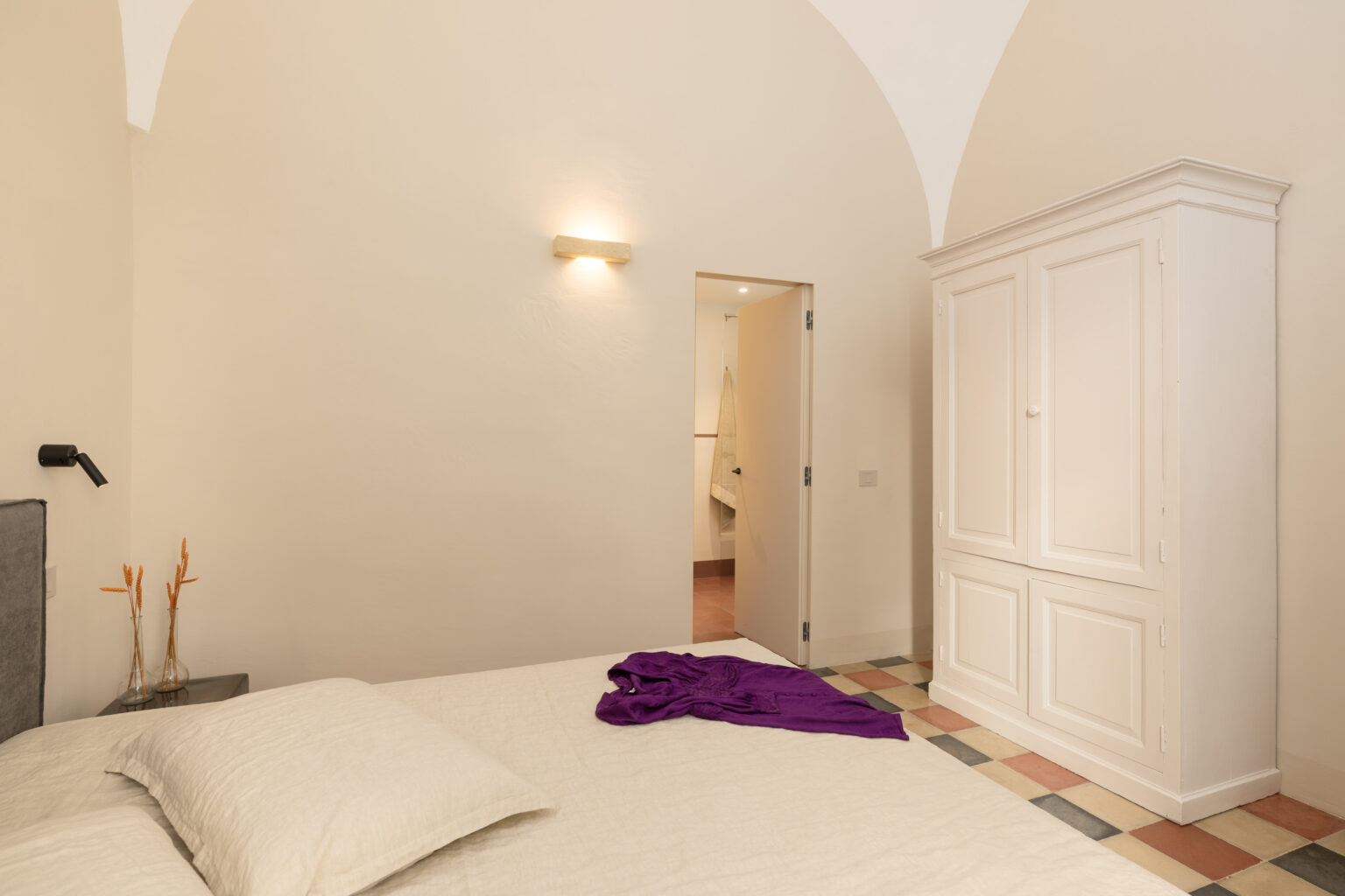 Interior view of a modern bedroom featuring a light-colored bed with white bedding, a purple garment draped over the bed, and a white wardrobe. The room has a warm ambiance with a wall-mounted light and an arched ceiling, complemented by a patterned tile floor. A doorway leads to an adjoining bathroom area. | Only on SopranoVillas