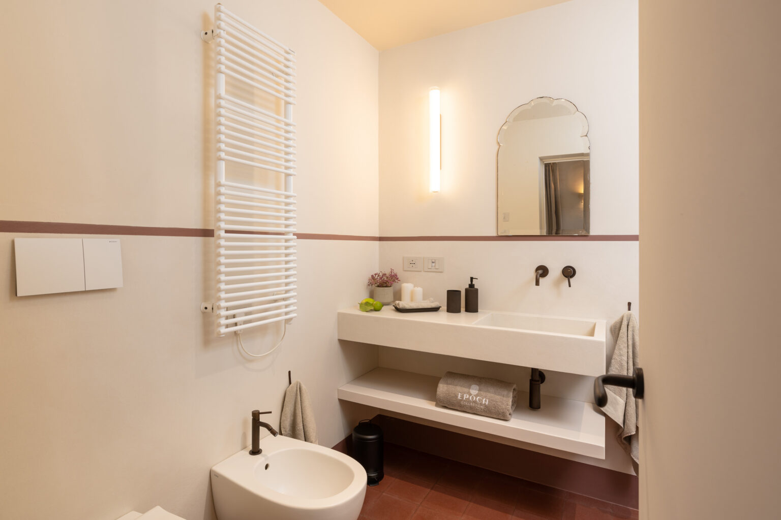 Modern bathroom interior featuring a minimalist design with a wall-mounted towel warmer, a sleek sink with modern fixtures, and a large mirror. The space includes neutral tones with a touch of color from decorative items, emphasizing functionality and style. | Only on SopranoVillas