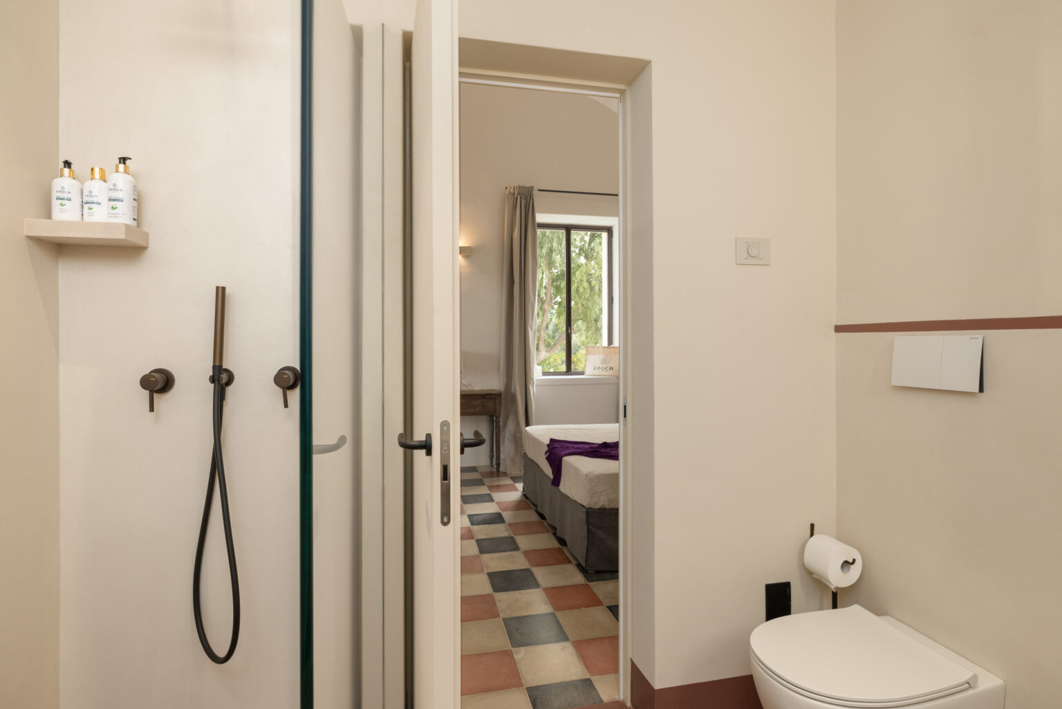 Modern bathroom interior featuring a shower, toilet, and colorful tiled floor, with a view into a well-lit bedroom beyond. | Only on SopranoVillas
