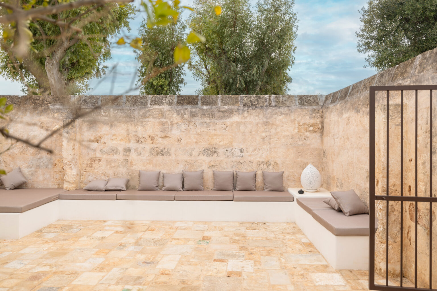 Stylish outdoor seating area with modern cushions, set against a textured stone wall, featuring potted trees and a decorative lantern. Ideal for relaxation and outdoor gatherings. | Only on SopranoVillas