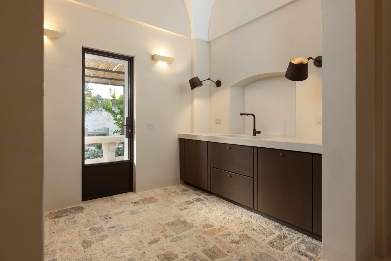 Modern kitchen interior featuring a minimalist design with dark cabinetry, a sleek countertop, and a stylish faucet. Natural light streams in through a door leading to an outdoor space, highlighting the stone floor and warm color palette. | Only on SopranoVillas