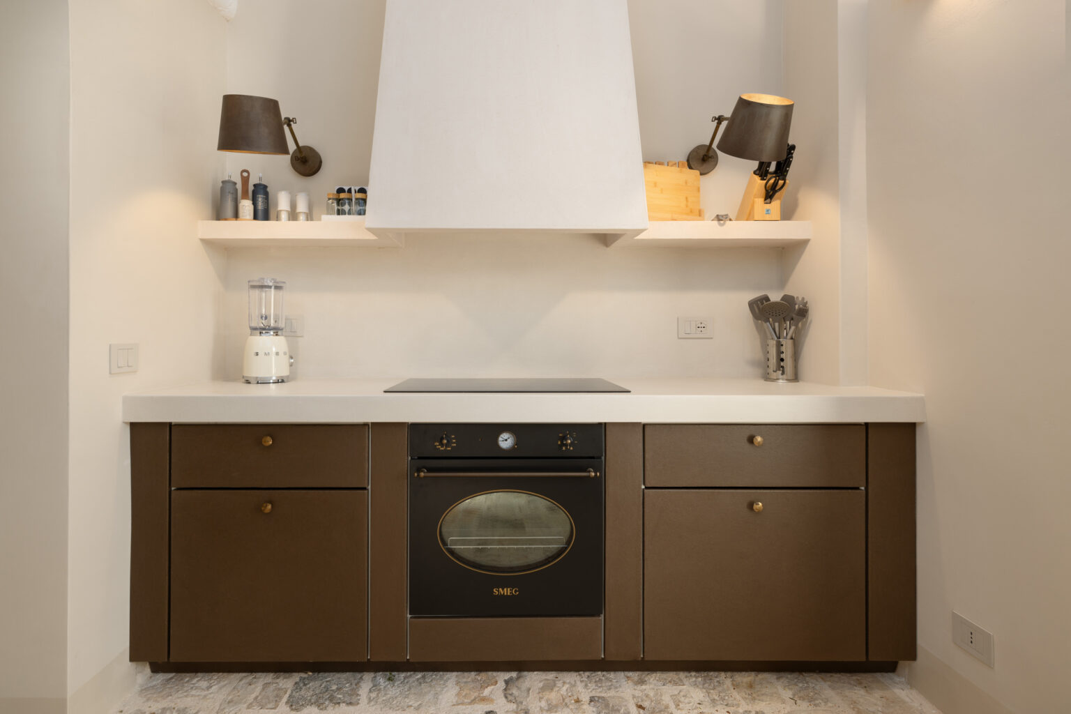 Modern kitchen design featuring a sleek SMEG oven, minimalist cabinetry in dark brown, a white countertop, and stylish wall-mounted lamps. The space includes a blender and kitchen utensils, providing a functional and aesthetic cooking environment. | Only on SopranoVillas