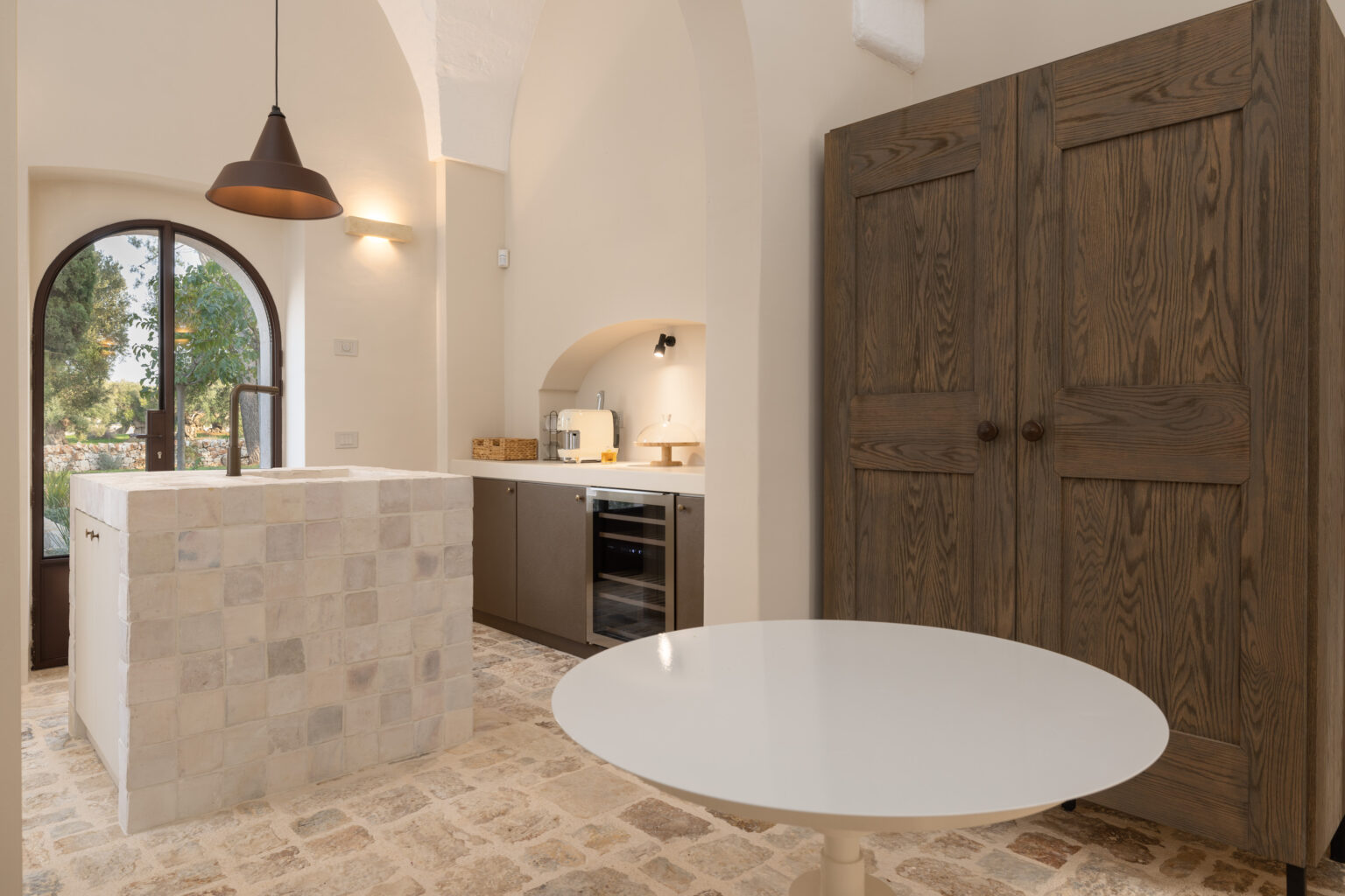 Modern kitchen interior featuring a stone island, sleek cabinetry, and a round dining table. Natural light streams through an arched window, highlighting the elegant design and earthy tones. Perfect for contemporary home decor inspiration. | Only on SopranoVillas