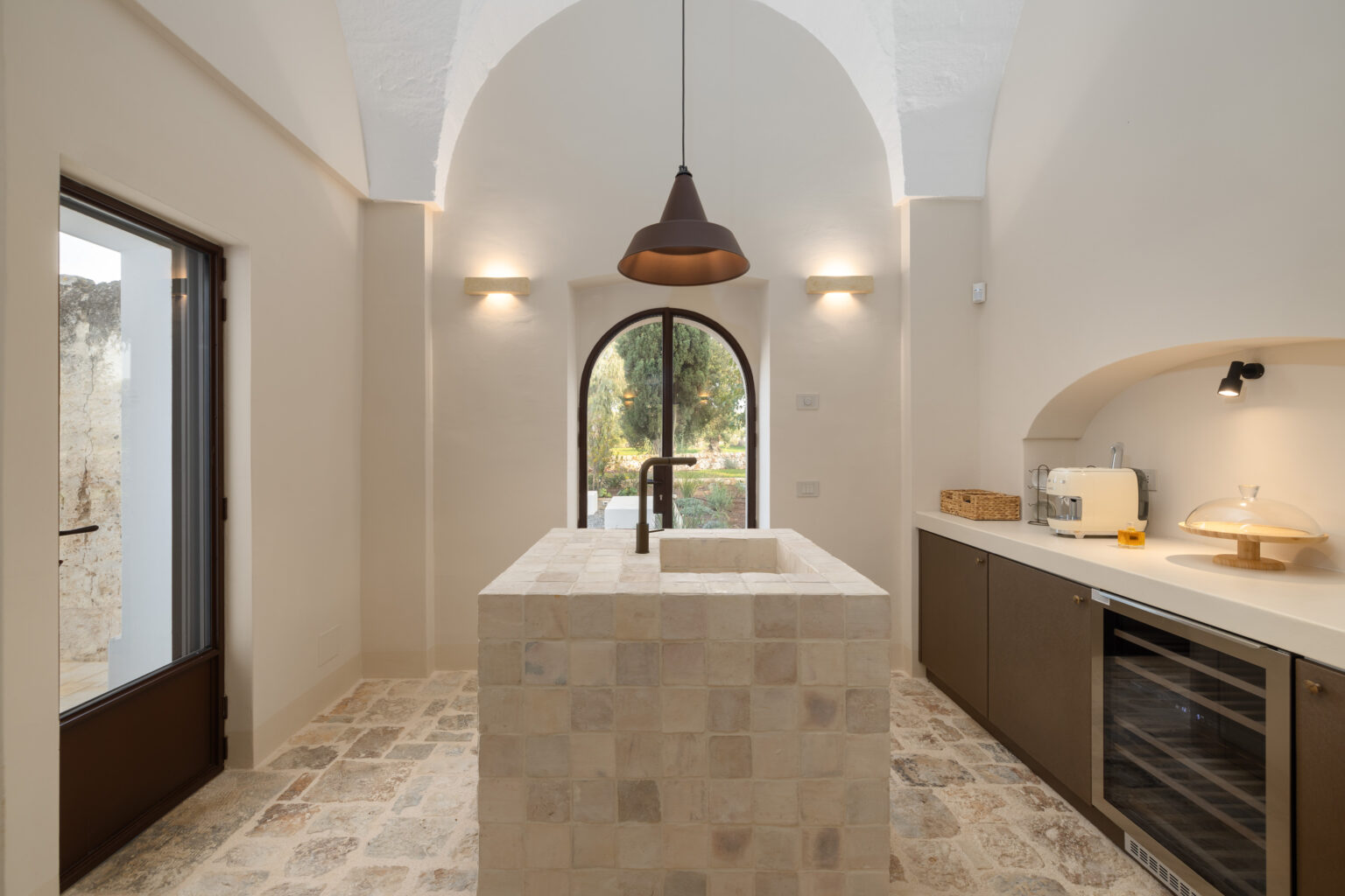Modern kitchen interior featuring a stone island, minimalist cabinetry, and large arched window. The design incorporates natural light, neutral tones, and stylish lighting fixtures, creating a warm and inviting atmosphere. | Only on SopranoVillas