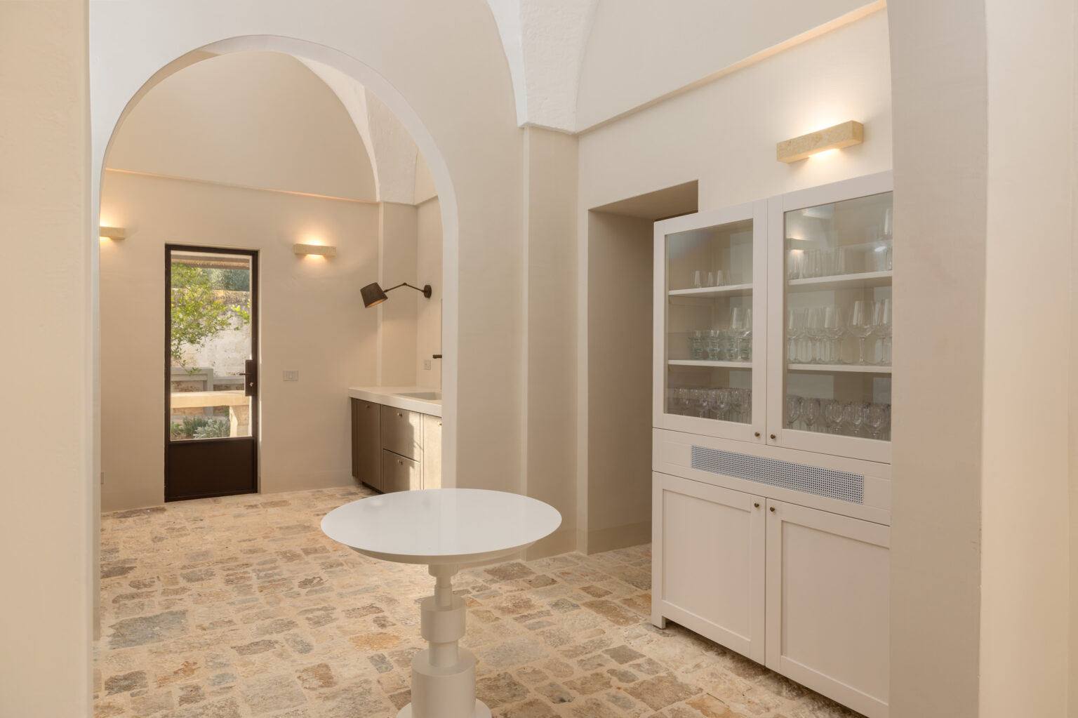 Modern kitchen interior featuring arched ceilings, a sleek countertop, a glass-front cabinet displaying glassware, and a round table. Natural light floods the space through a door leading to an outdoor area, highlighting the rustic stone flooring. | Only on SopranoVillas