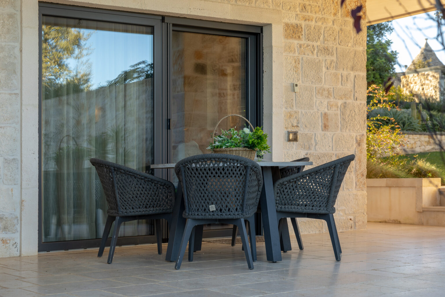 Outdoor dining area featuring a modern black wicker table and four matching chairs, set against a backdrop of a stone wall and glass sliding doors. | Only on SopranoVillas