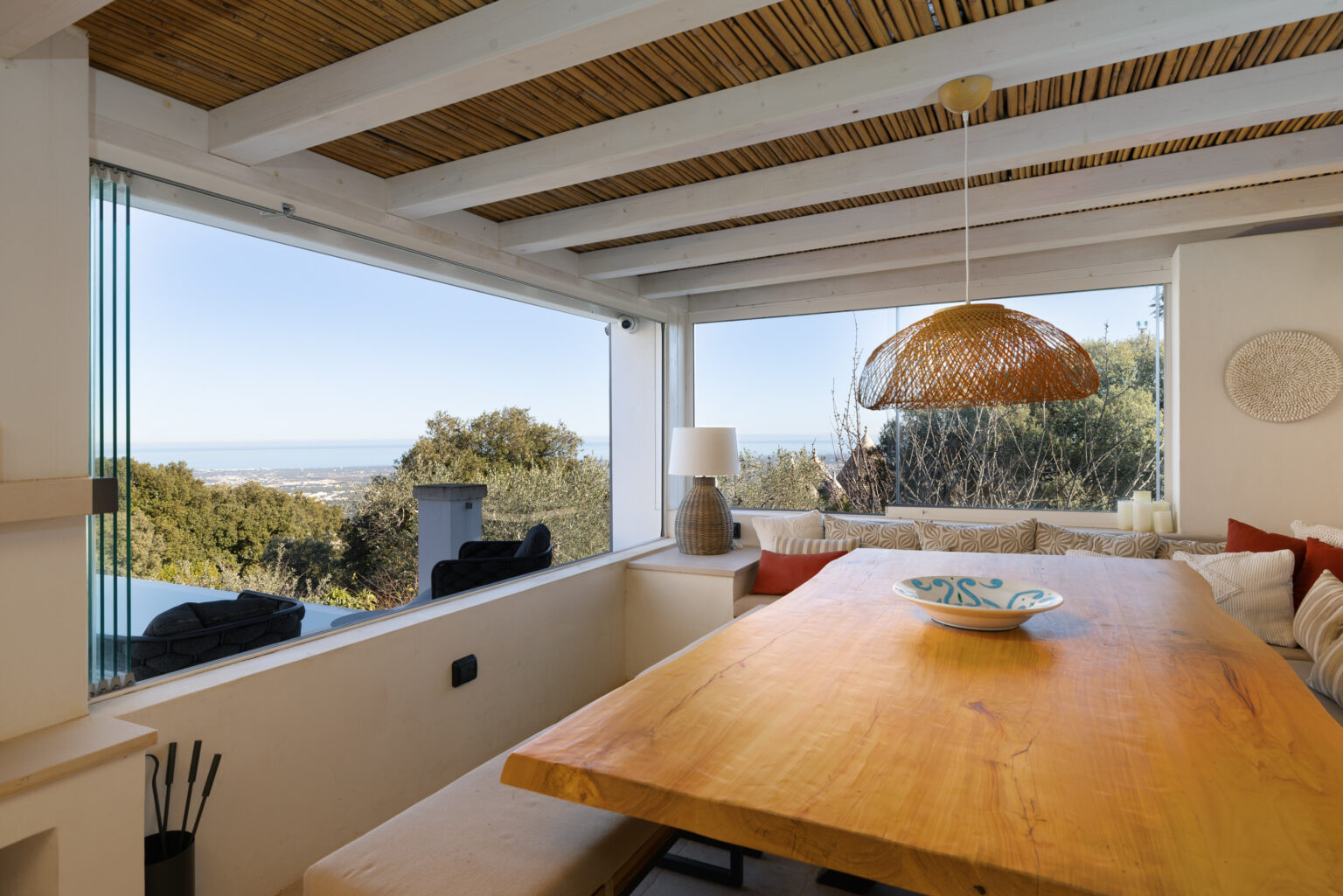 Modern dining area with a wooden table, large windows showcasing a scenic view, and a stylish pendant light. The space features a cozy seating area with decorative pillows, blending natural elements with contemporary design. | Only on SopranoVillas
