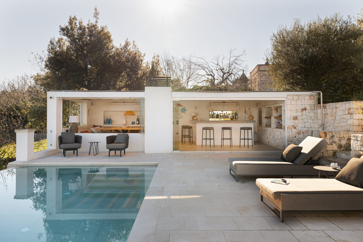 Modern outdoor living space featuring a stylish pool, lounge chairs, and an open-concept kitchen area with seating. Surrounded by greenery, this serene setting is perfect for relaxation and entertaining. | Only on SopranoVillas