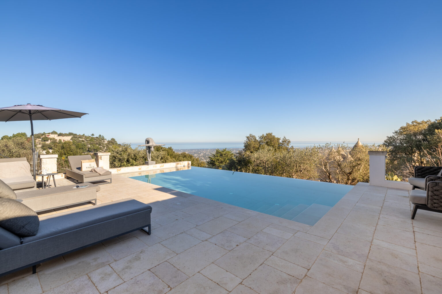 Luxury outdoor pool area with modern lounge chairs, an umbrella, and stunning views of the landscape under a clear blue sky. | Only on SopranoVillas
