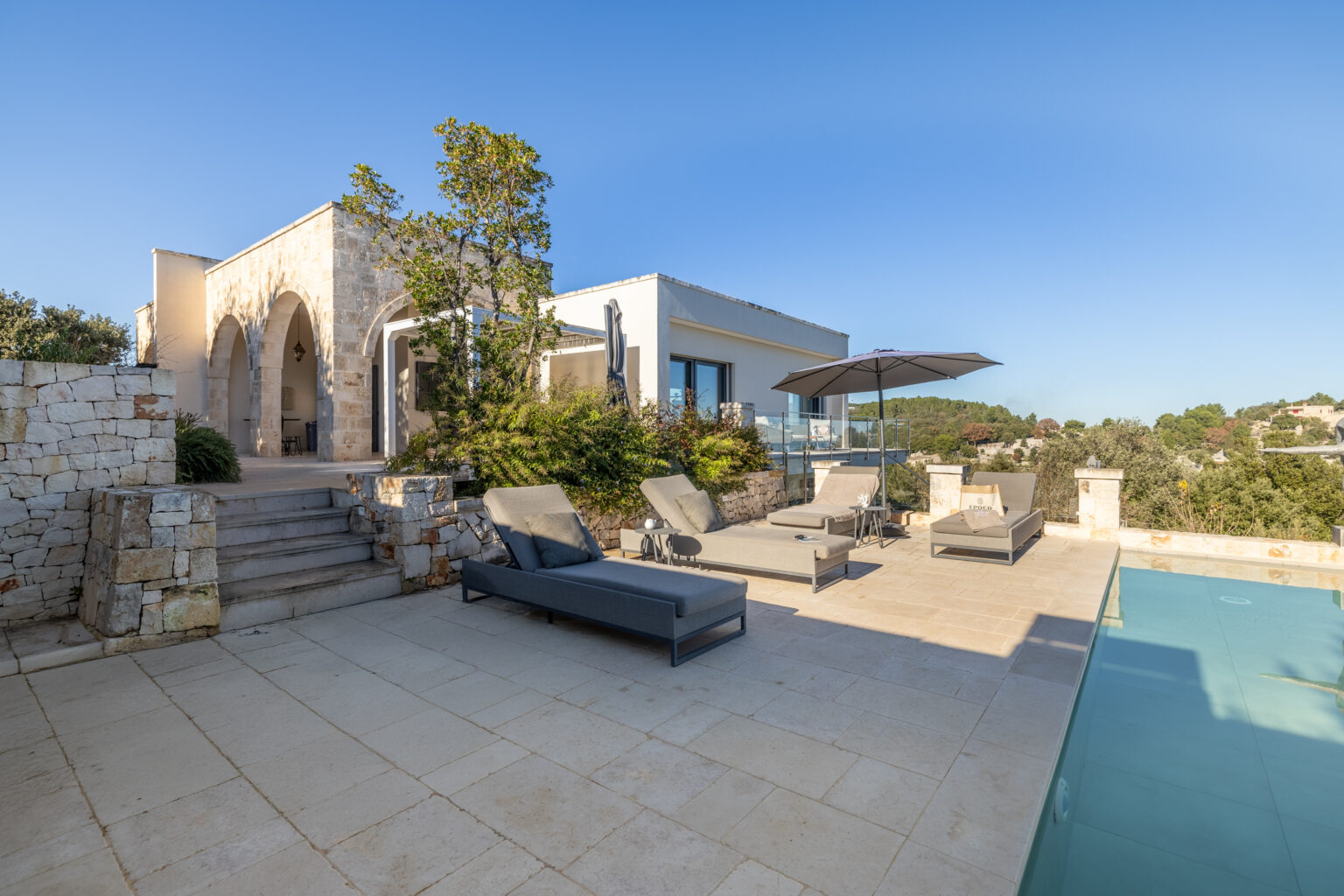 Modern luxury villa with a spacious outdoor area, featuring a pool, sun loungers, and lush greenery, set against a clear blue sky. | Only on SopranoVillas