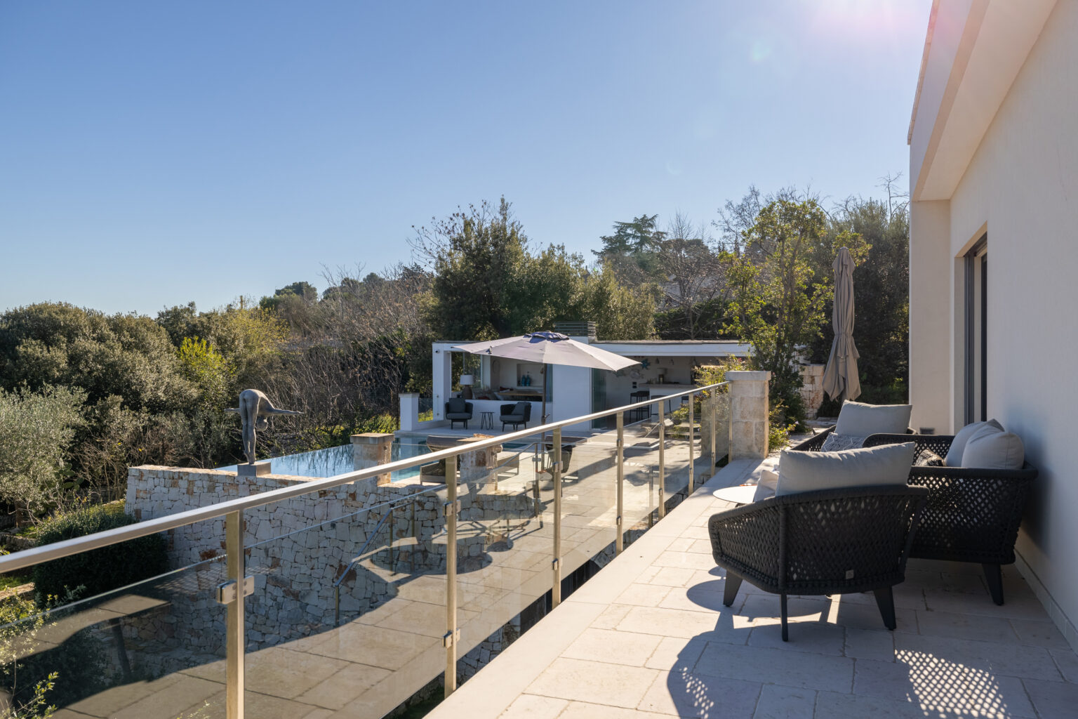 Modern villa terrace overlooking a serene landscape with a swimming pool, outdoor seating area, and lush greenery under clear blue skies. Perfect for relaxation and outdoor entertaining. | Only on SopranoVillas